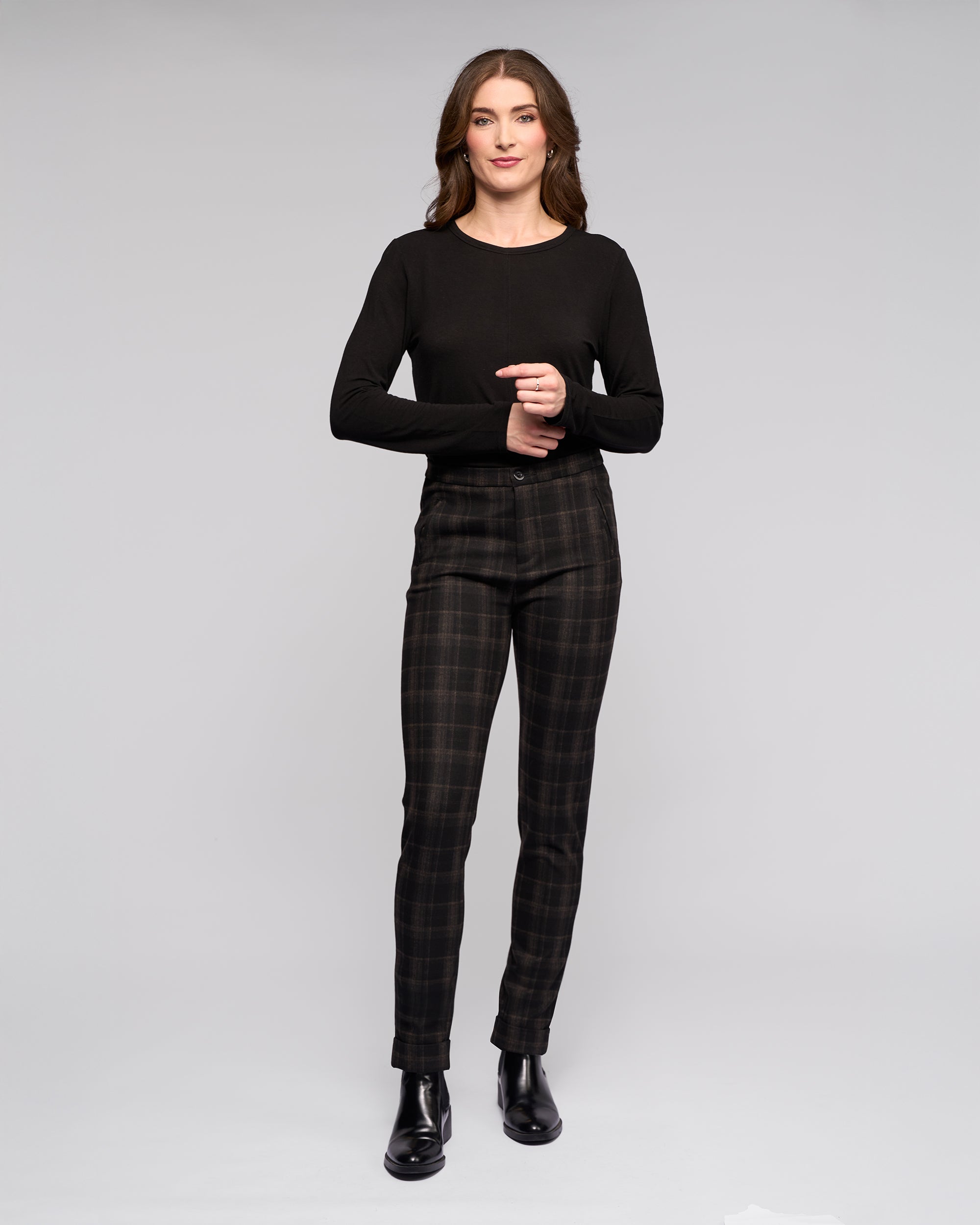 A woman with loose brown hair smiles slightly, clasping her hands at waist height. She wears a black long-sleeve top, Vassalli Style Pant - Checkmate, and black ankle boots against a plain light gray background.