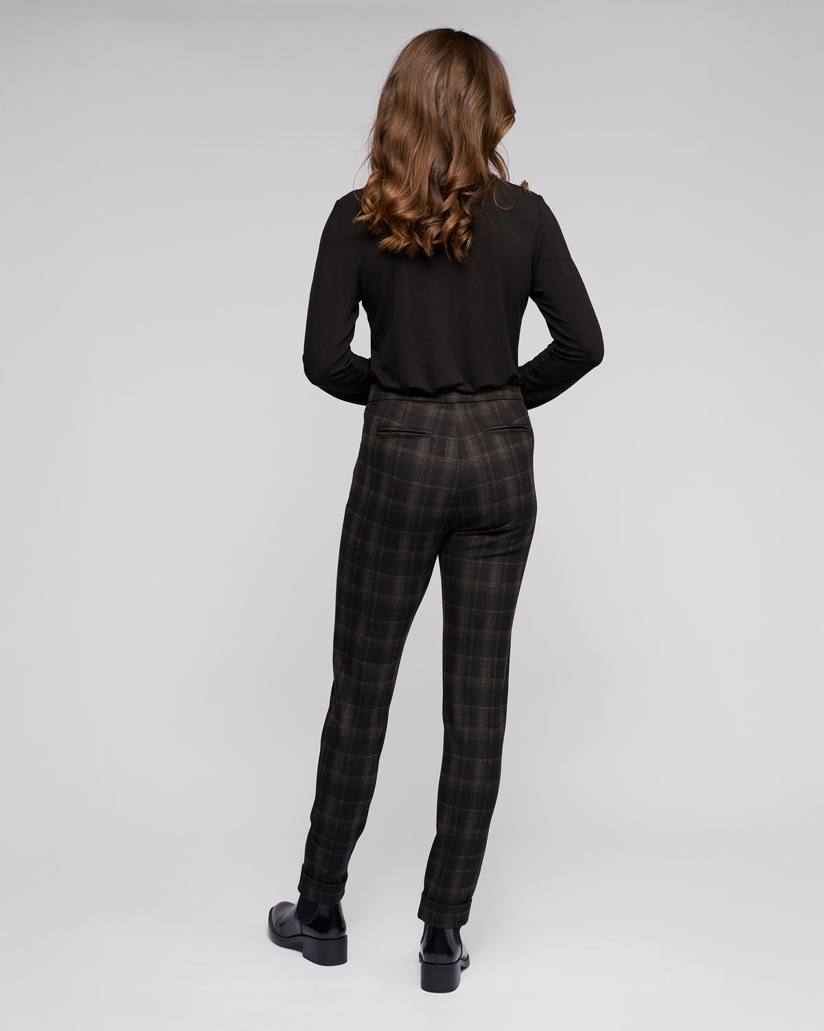 A person with wavy brown hair stands facing away, wearing a long-sleeved black top, Vassalli Style Pant - Checkmate, and black shoes against a light gray backdrop.