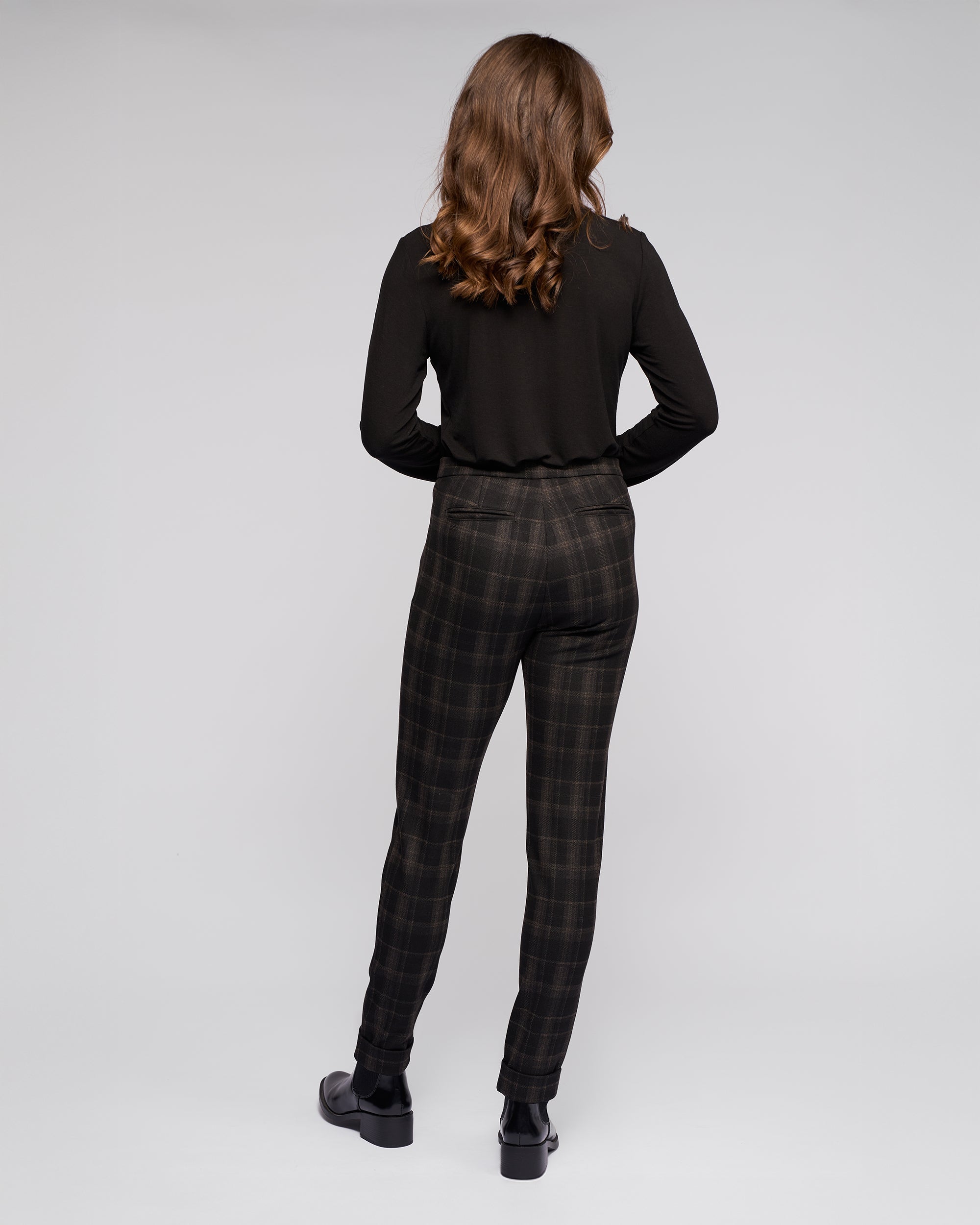 A person with wavy brown hair stands facing away, wearing a long-sleeved black top, Vassalli Style Pant - Checkmate, and black shoes against a light gray backdrop.