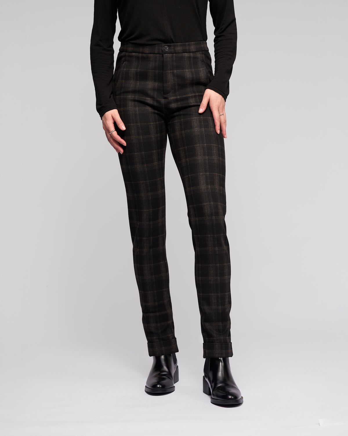 A person models Vassalli Style Pant - Checkmate with skinny legs, paired with a black long-sleeve top and black ankle boots against a plain light gray background. Only their body from shoulders down is visible, hands relaxed by their sides.