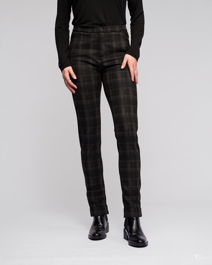 A person models Vassalli Style Pant - Checkmate with skinny legs, paired with a black long-sleeve top and black ankle boots against a plain light gray background. Only their body from shoulders down is visible, hands relaxed by their sides.