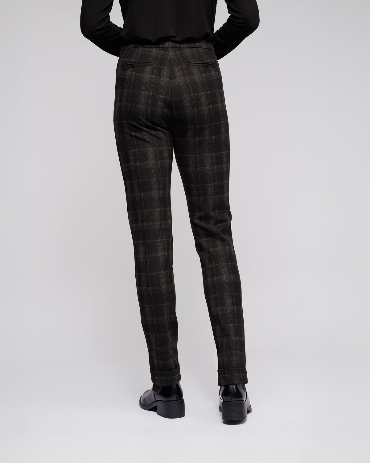 A person faces away from the camera, wearing Vassalli's Style Pant - Checkmate in black and brown check, paired with a black top and black shoes, against a plain light background.