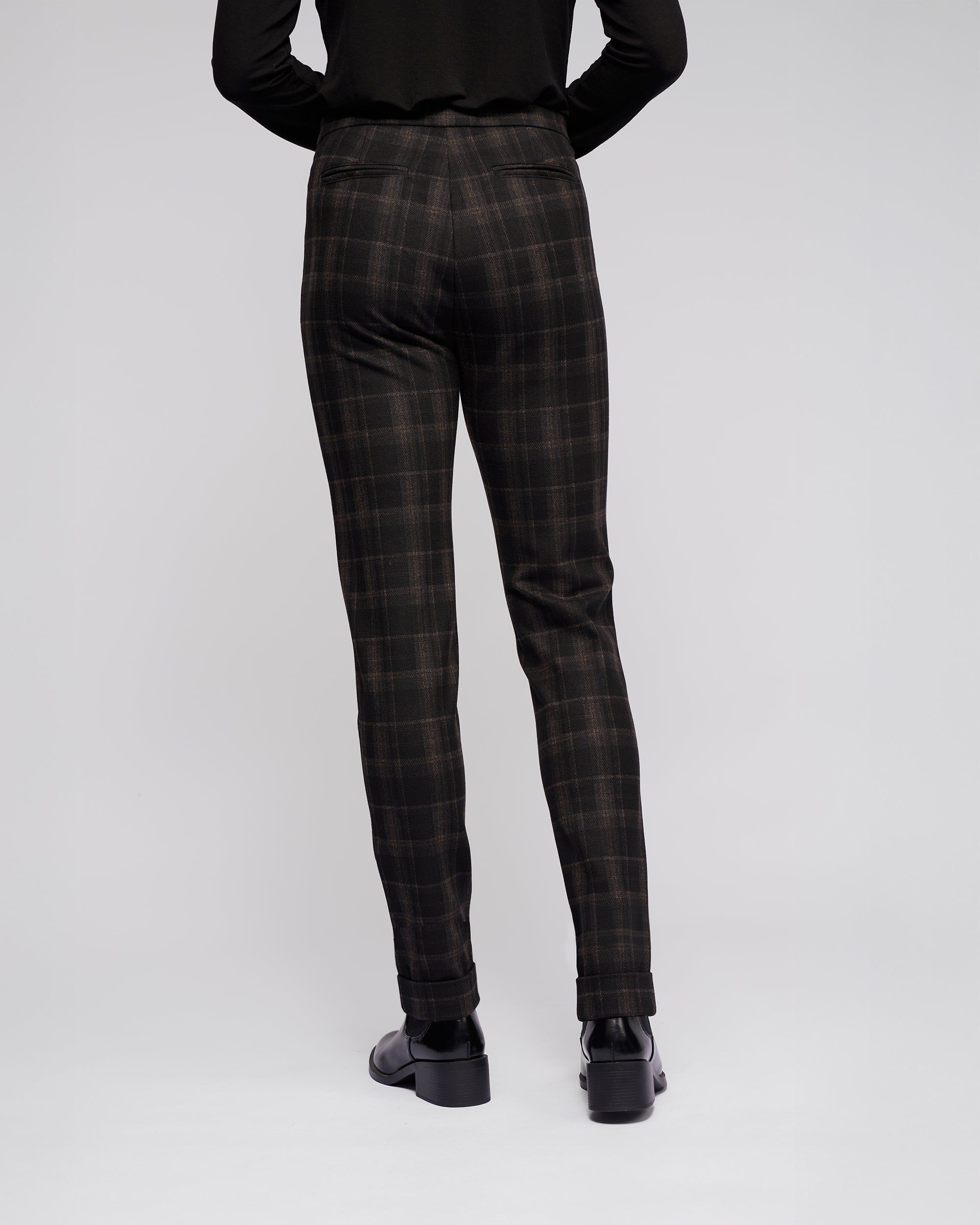 A person faces away from the camera, wearing Vassalli's Style Pant - Checkmate in black and brown check, paired with a black top and black shoes, against a plain light background.