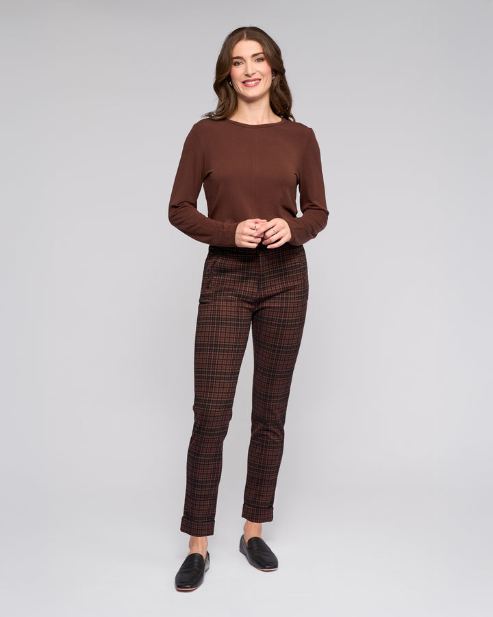 A woman with long brown hair smiles against a gray background, wearing the Vassalli Style Pant - Cherry and a brown stretch knit top. Her hands are gently clasped in front of her.