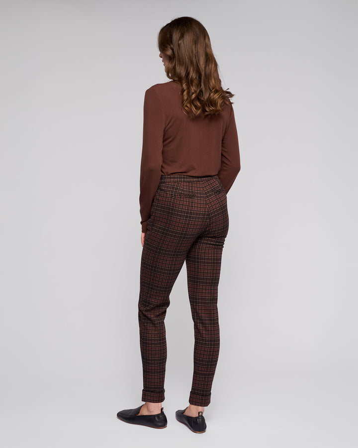 A woman stands facing away, wearing a long-sleeved brown top, Vassalli Style Pant - Cherry in stretch knit fabric (skinny-leg), and black slip-on shoes, set against a plain light gray background.