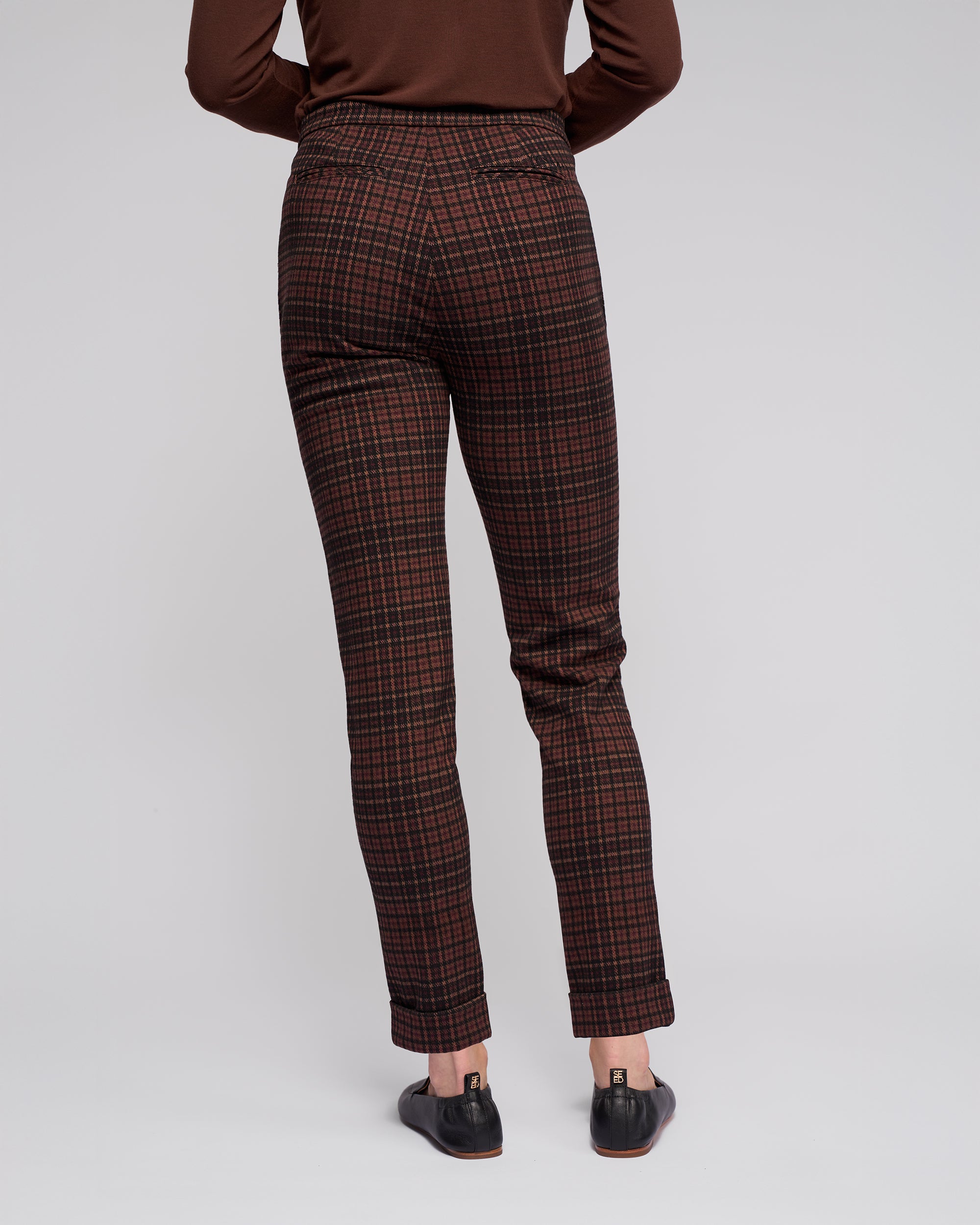 A person stands facing away, wearing Vassalli's Style Pant - Cherry in slim-fit check plaid stretch knit, paired with a brown long-sleeve top and black shoes against a plain light background.