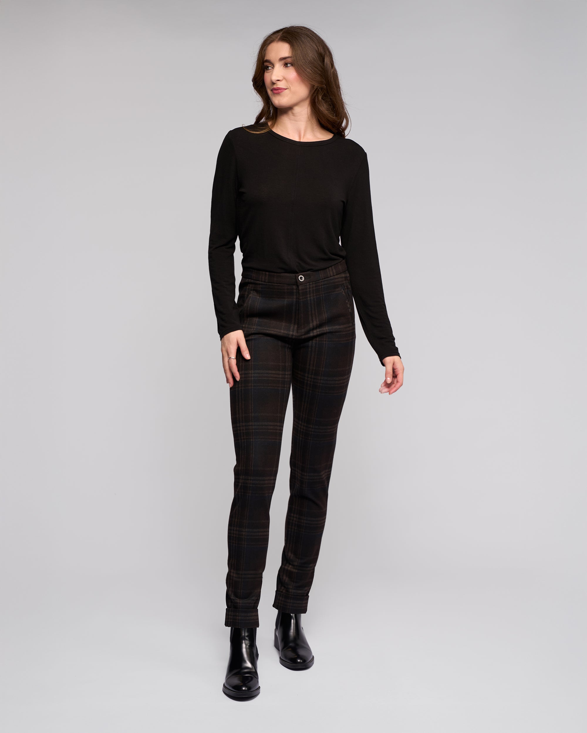 A woman stands against a plain gray background, wearing a long-sleeve black top and Vassalli Style Pant - Hugo in dark plaid. She pairs the look with black ankle boots, her long brown hair framing her relaxed expression as she gazes to the side.