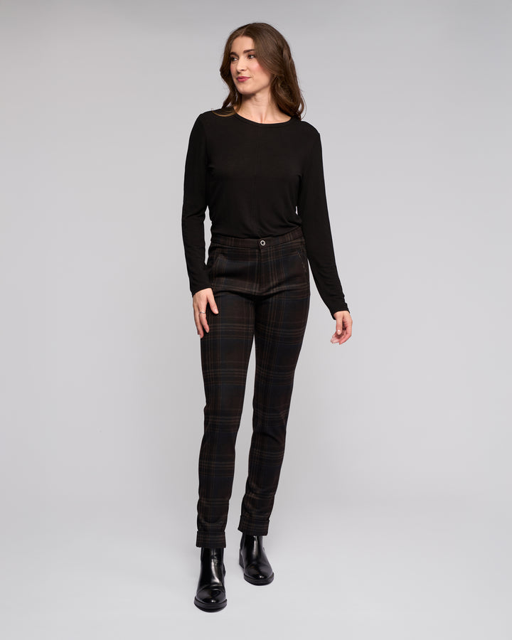 A woman stands against a plain gray background, wearing a long-sleeve black top and Vassalli Style Pant - Hugo in dark plaid. She pairs the look with black ankle boots, her long brown hair framing her relaxed expression as she gazes to the side.
