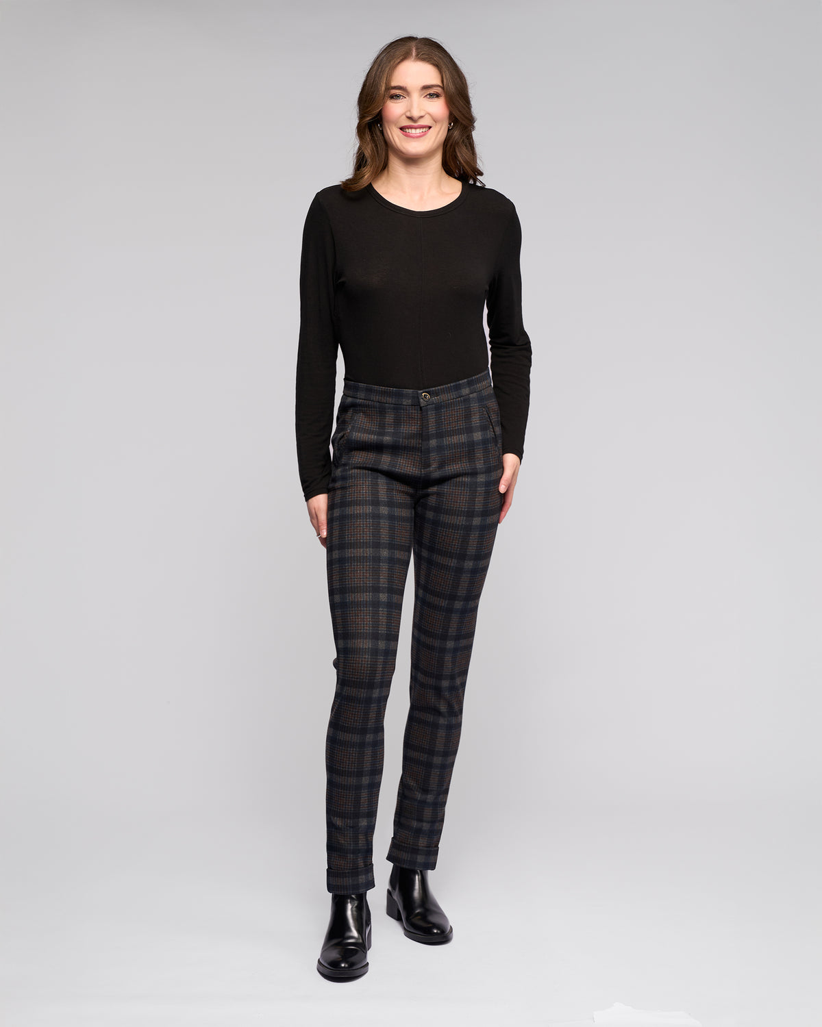 A woman wears Vassalli's Style Pant - Nomad in navy and brown check with a black long-sleeve top and black ankle boots, standing against a light grey background.