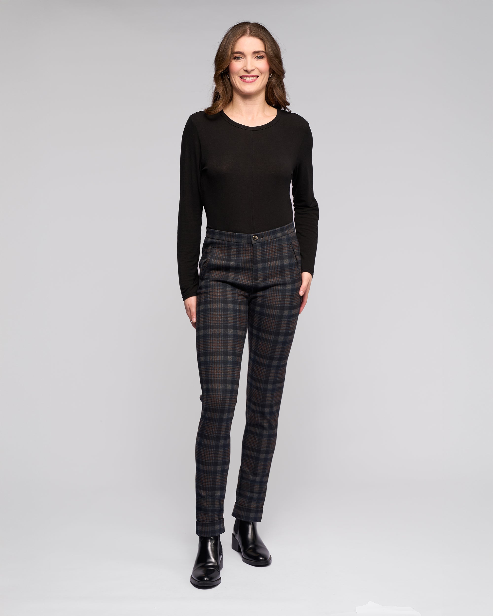 A woman wears Vassalli's Style Pant - Nomad in navy and brown check with a black long-sleeve top and black ankle boots, standing against a light grey background.
