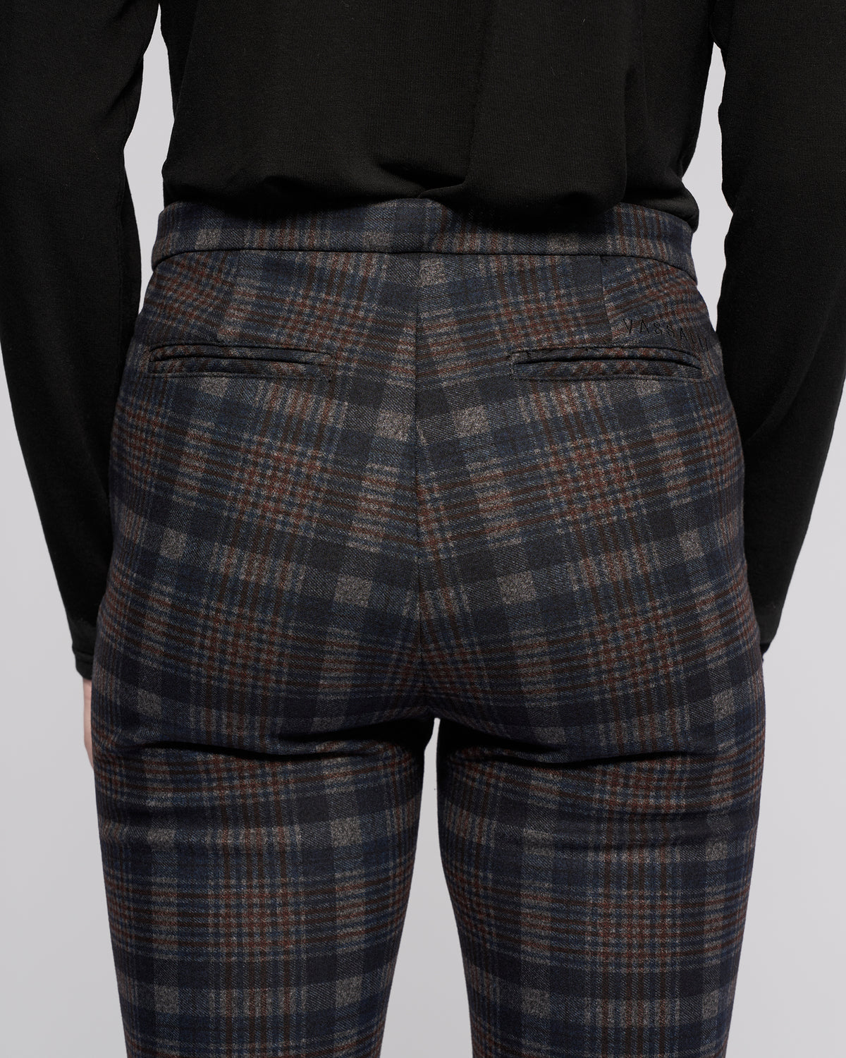 A person is seen from the back wearing Vassalli Style Pant - Nomad, featuring a blue, gray, and brown Nomad check plaid pattern with two narrow back pockets, paired with a black long-sleeve top.