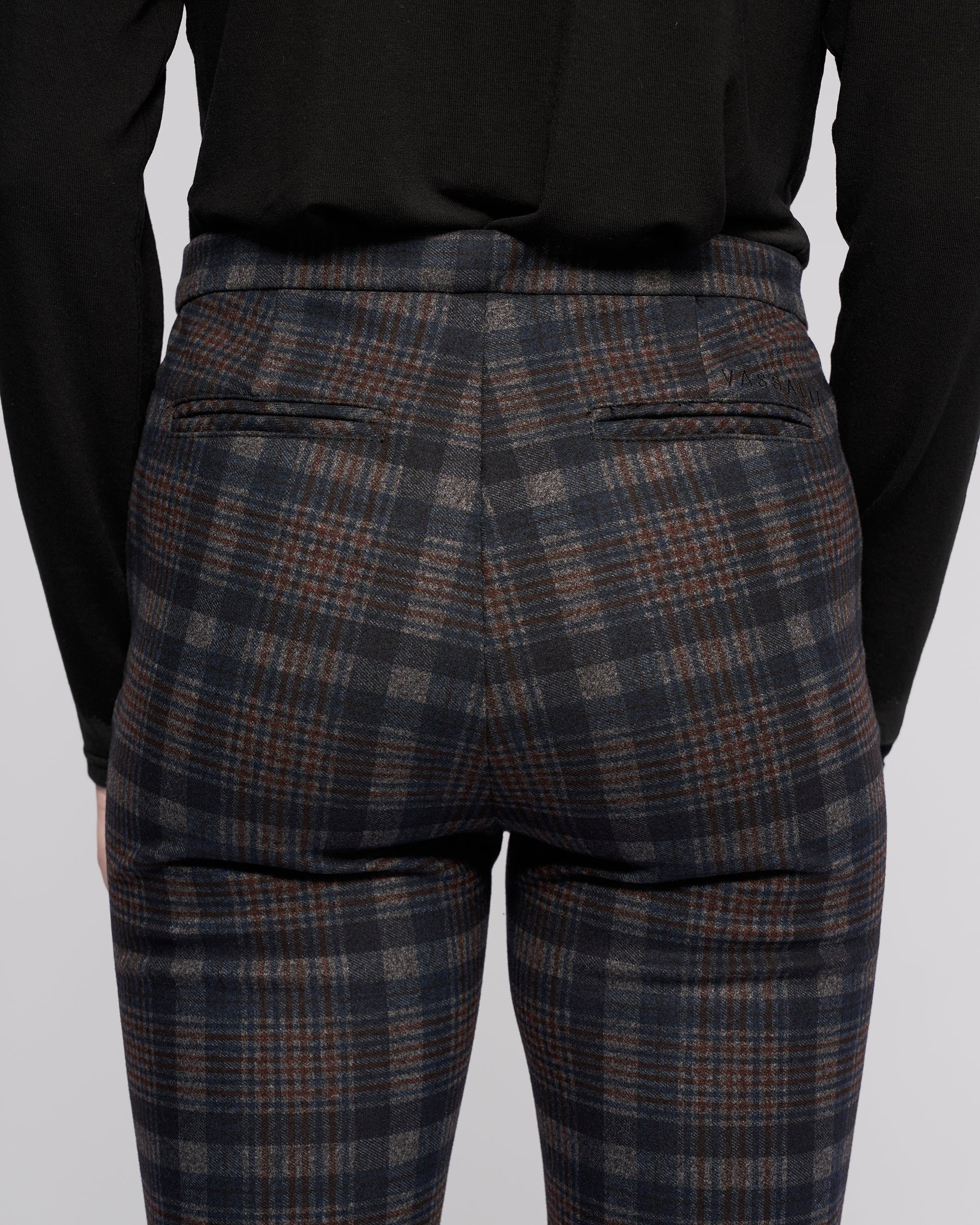 A person is seen from the back wearing Vassalli Style Pant - Nomad, featuring a blue, gray, and brown Nomad check plaid pattern with two narrow back pockets, paired with a black long-sleeve top.