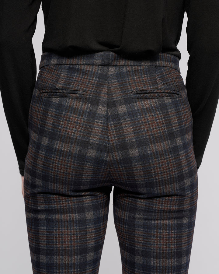 A person is seen from the back wearing Vassalli Style Pant - Nomad, featuring a blue, gray, and brown Nomad check plaid pattern with two narrow back pockets, paired with a black long-sleeve top.