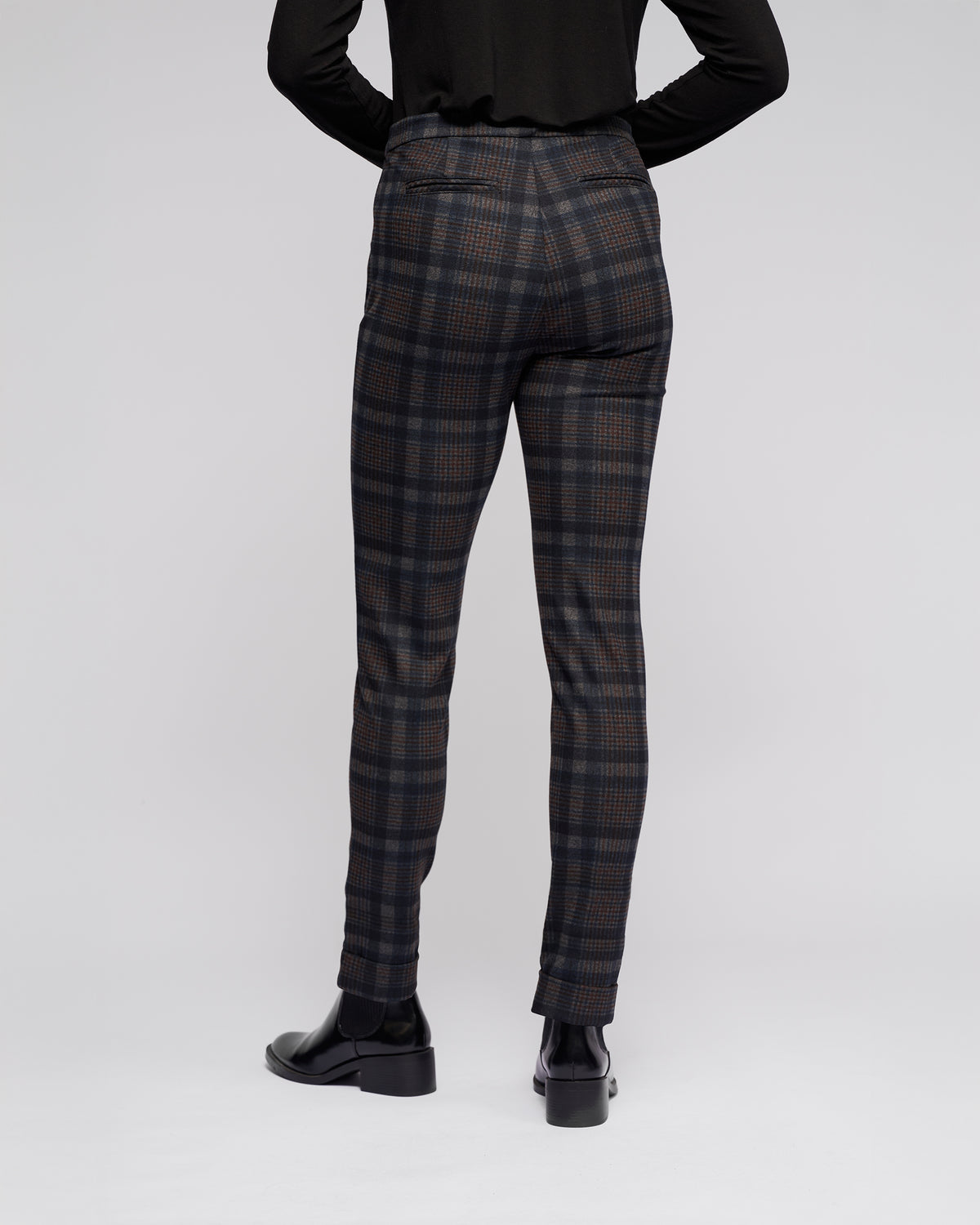 A person stands facing away, wearing Vassalli Style Pant - Nomad, a black long-sleeve top, and black heeled shoes against a plain light background.