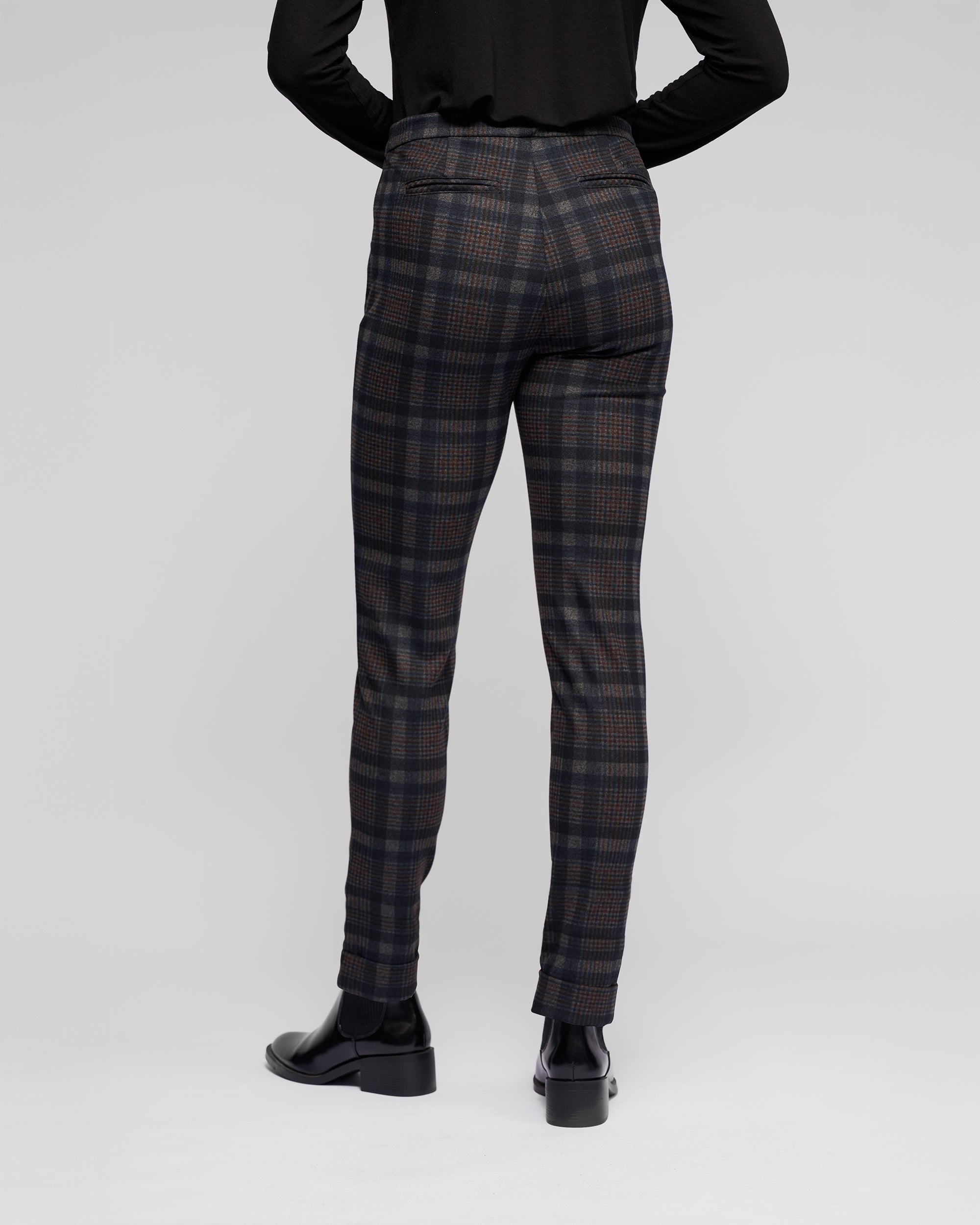 A person stands facing away, wearing Vassalli Style Pant - Nomad, a black long-sleeve top, and black heeled shoes against a plain light background.