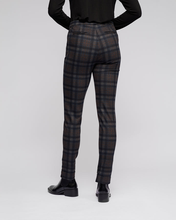 A person stands facing away, wearing Vassalli Style Pant - Nomad, a black long-sleeve top, and black heeled shoes against a plain light background.