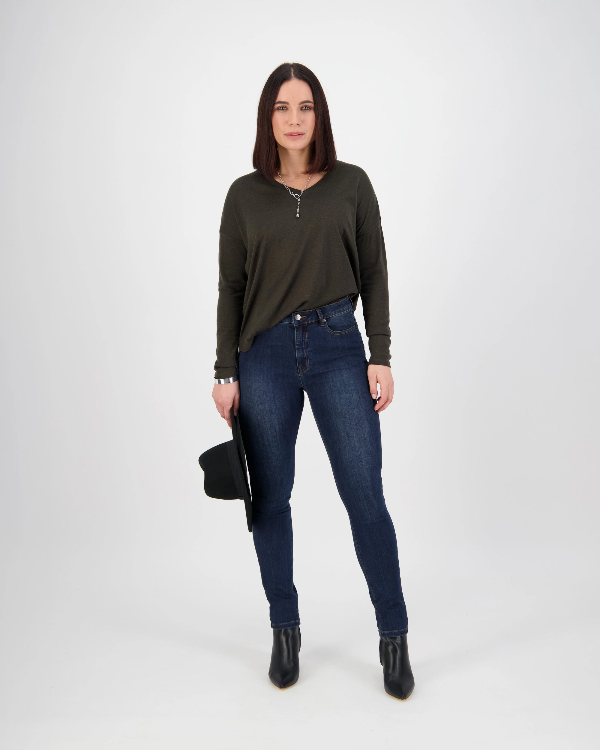 A woman with straight brown hair stands against a white background wearing Vassalli’s Notion Jean in Indigo Denim, a dark green long-sleeve top, and black ankle boots. She holds a black hat in her left hand and looks neutrally at the camera.