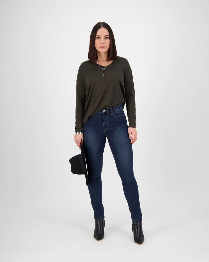 A woman with straight brown hair stands against a white background wearing Vassalli’s Notion Jean in Indigo Denim, a dark green long-sleeve top, and black ankle boots. She holds a black hat in her left hand and looks neutrally at the camera.