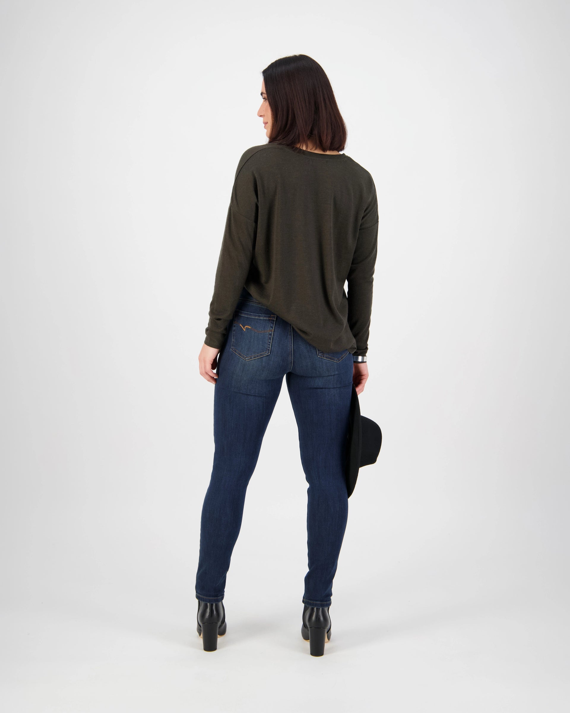 A woman faces away in a Vassalli Notion Jean - Indigo Denim, a dark long-sleeve top, black heeled boots, and holds a black hat in her left hand against a plain light background.