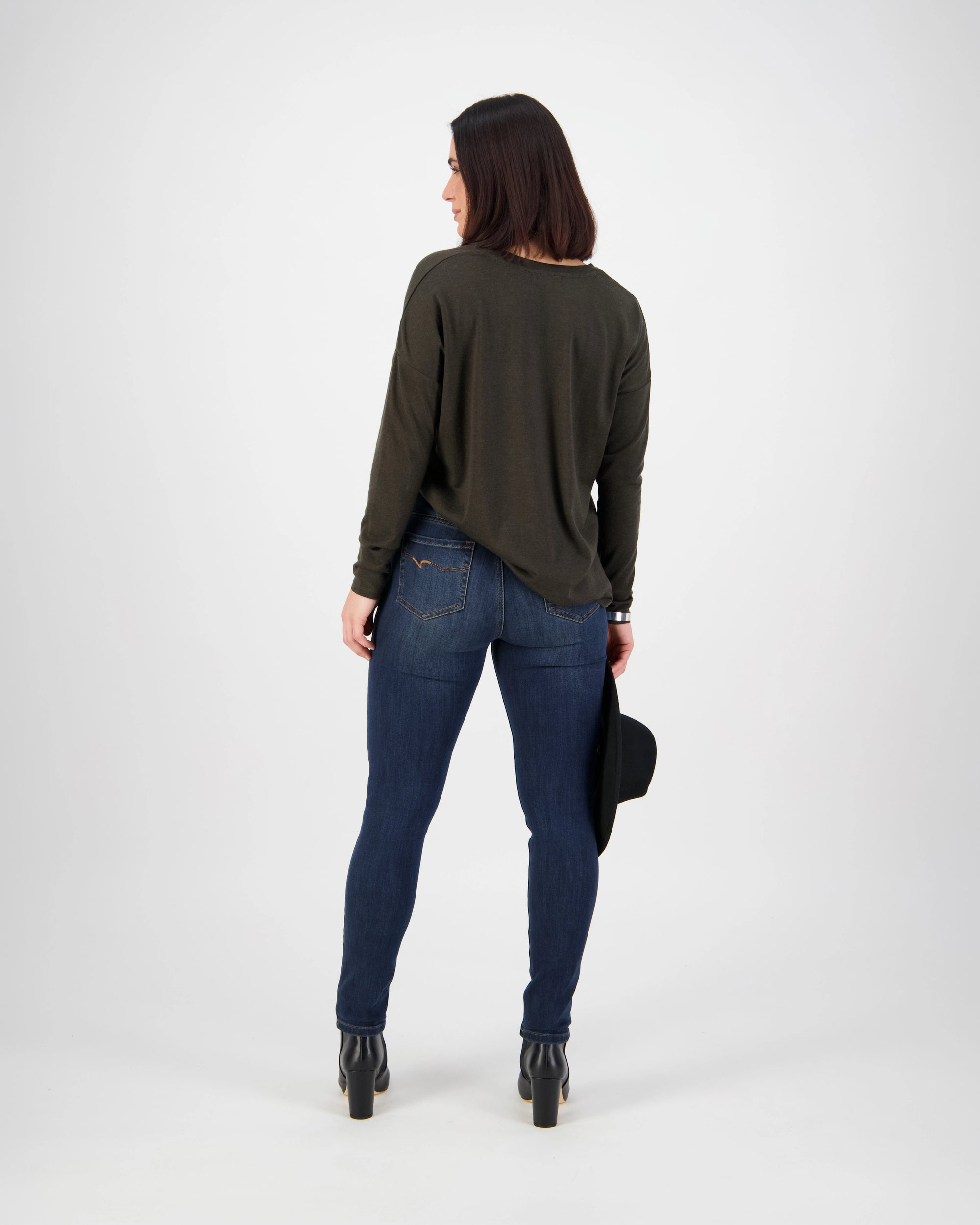 A woman faces away in a Vassalli Notion Jean - Indigo Denim, a dark long-sleeve top, black heeled boots, and holds a black hat in her left hand against a plain light background.