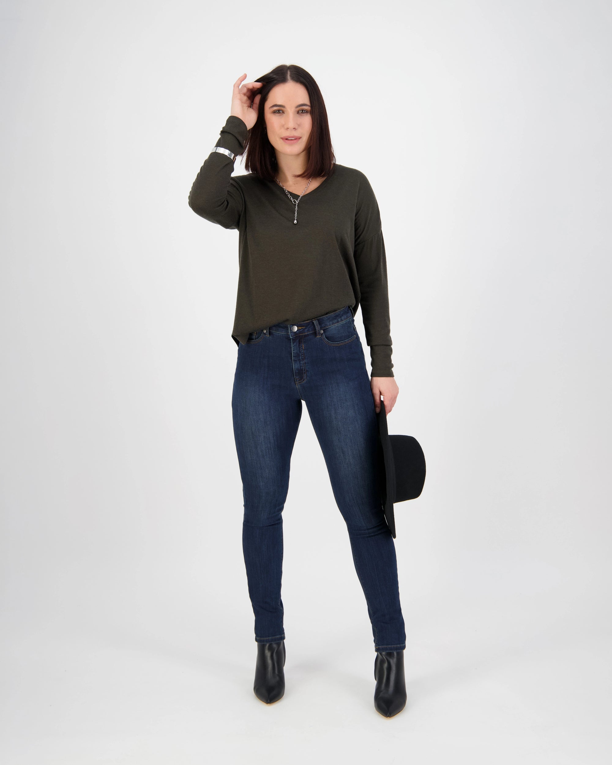 A woman with straight dark hair stands against a white background in a dark green long-sleeve top and Vassalli Notion Jean - Indigo Denim, paired with black ankle boots. She holds a black hat in her left hand and touches her head with her right.