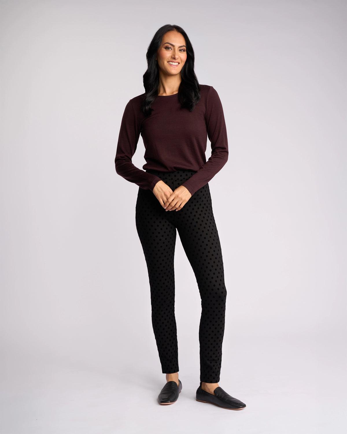 A woman with long dark hair smiles, wearing a maroon long-sleeve top, black Vassalli View Pant in a skinny fit, and black slip-on shoes, standing against a plain light background.