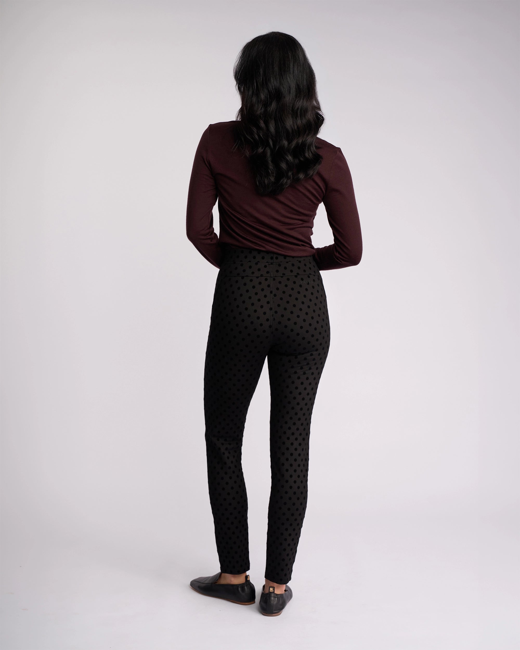 Wearing the Vassalli View Pant - Black, a person with long dark hair and a maroon long-sleeve top stands with their back to the camera against a plain light background.