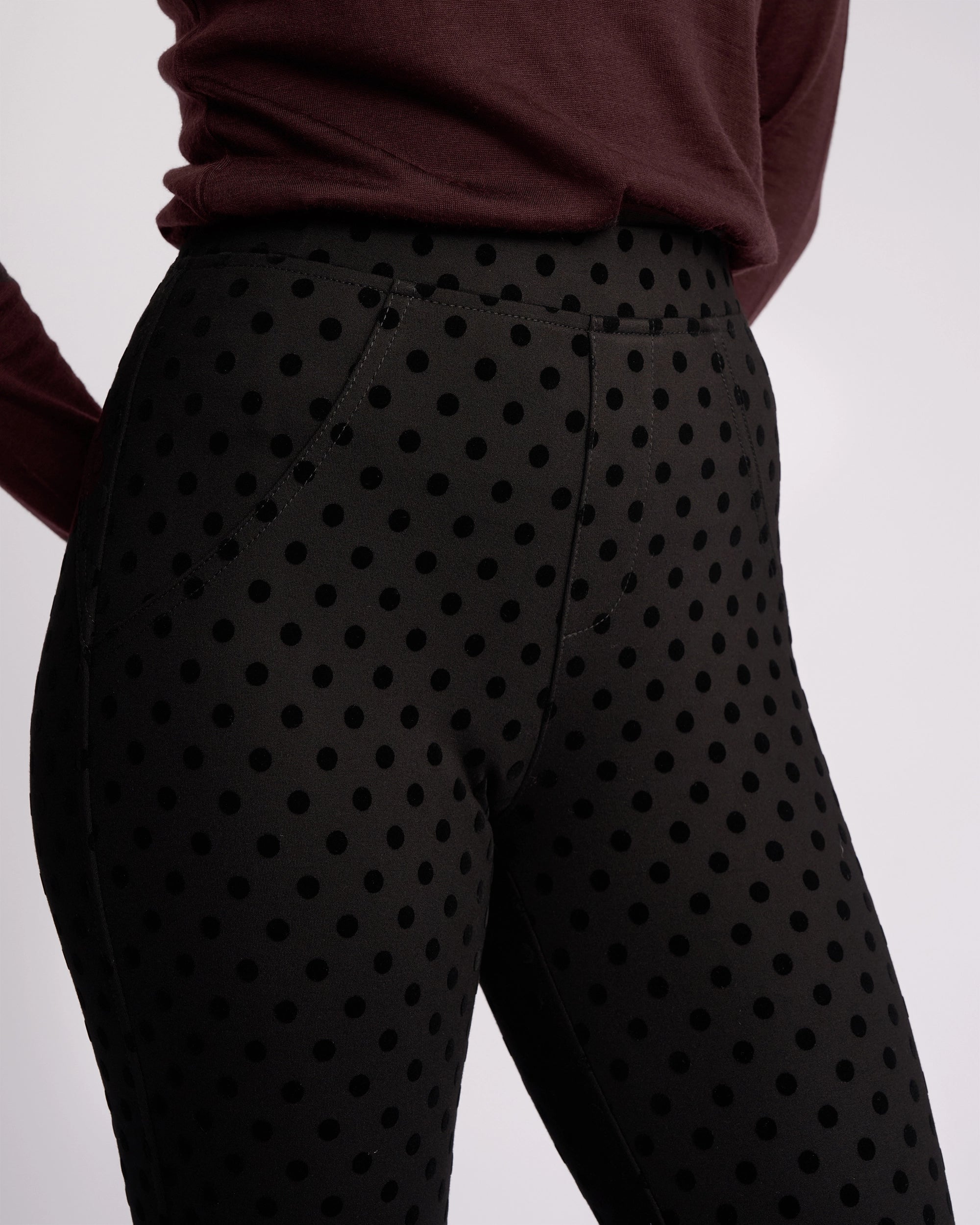 A person wearing Vassalli’s View Pant - Black, ankle-grazer high-waisted pants with large black polka dots, paired with a long-sleeved maroon top, is photographed from the waist down against a plain background.