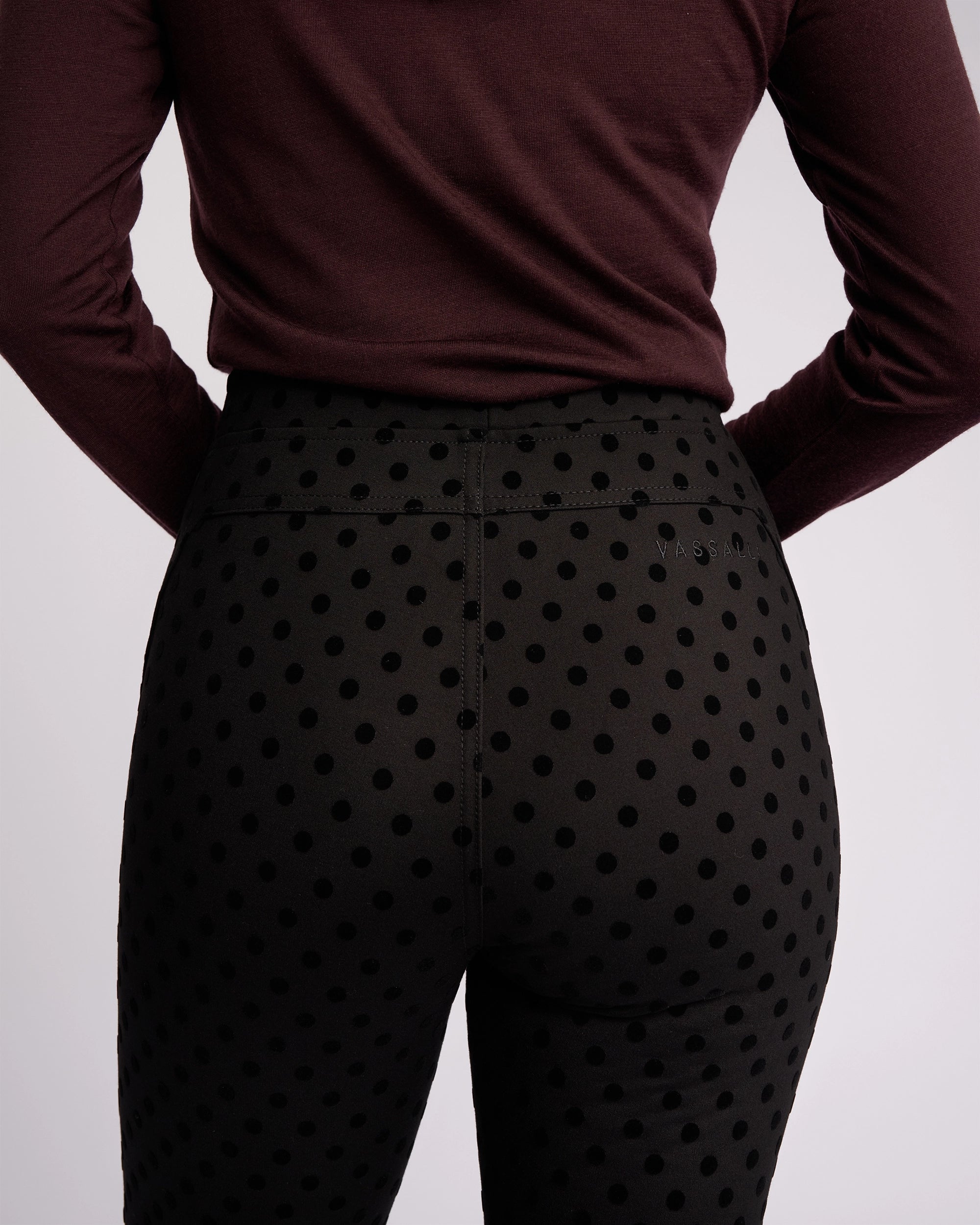 A person is shown from the back against a plain light background, wearing Vassalli’s View Pant - Black, a high-waisted skinny-leg pant in ponti fabric with black velvet polka dots, paired with a long-sleeved maroon top.