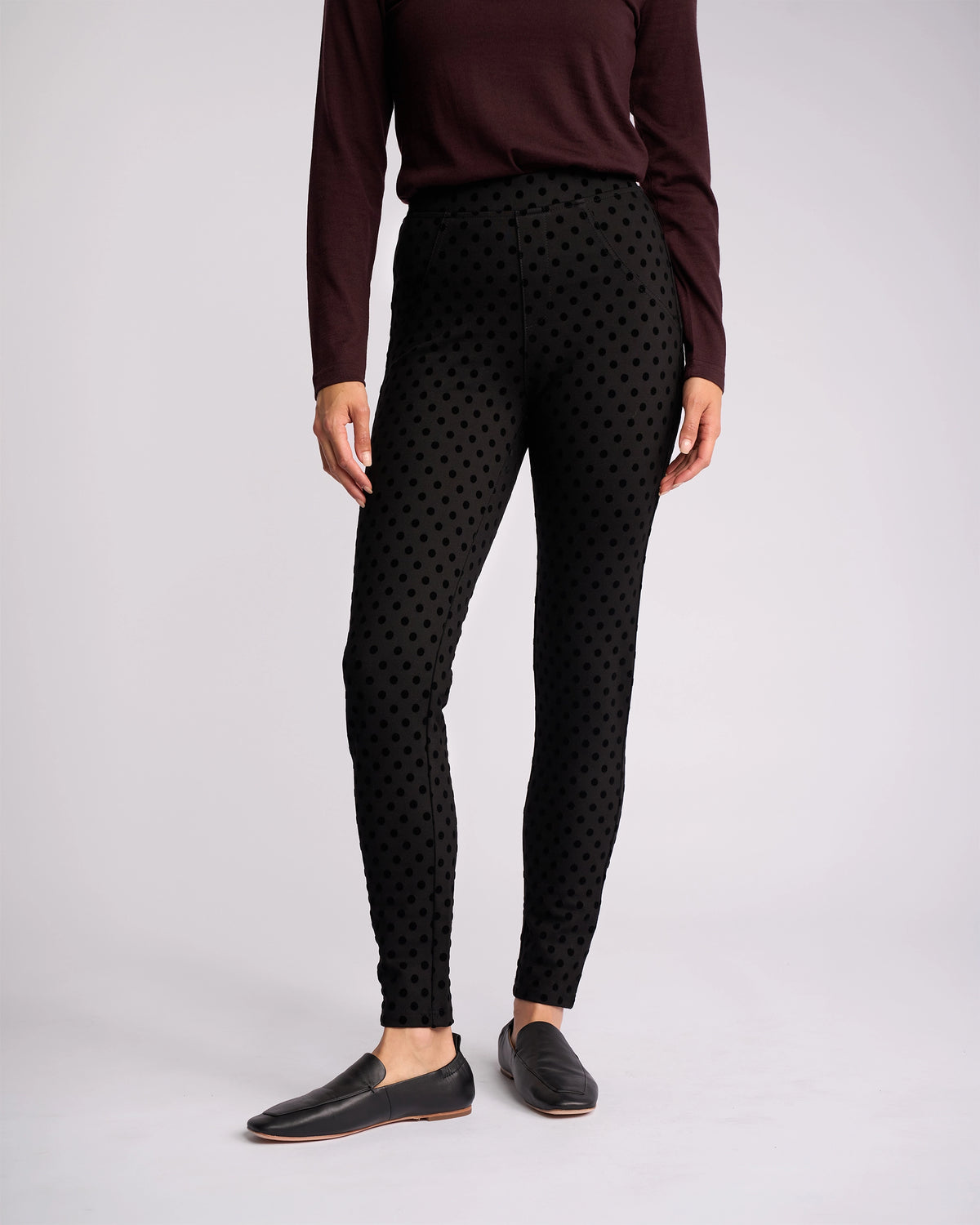 A person models the Vassalli View Pant - Black paired with a long-sleeve maroon top and black slip-on shoes, standing against a plain light background. The individual’s head is not visible in the image.
