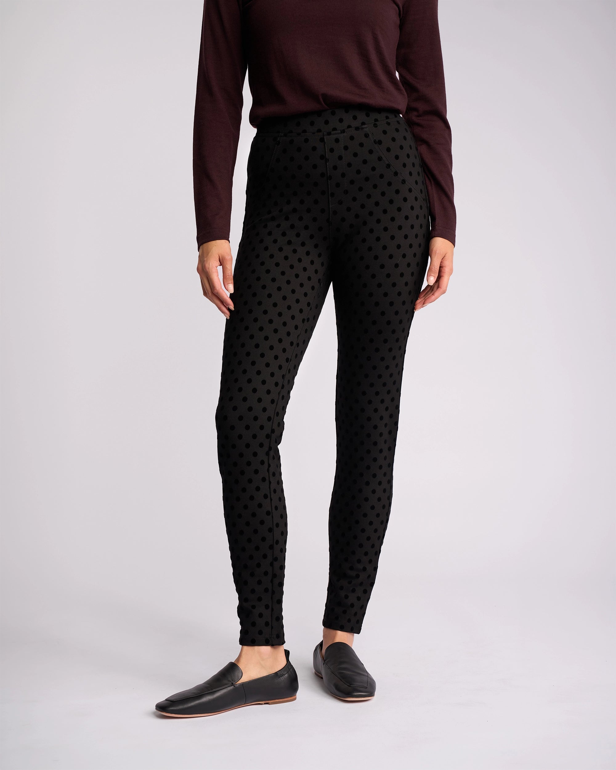 A person models the Vassalli View Pant - Black paired with a long-sleeve maroon top and black slip-on shoes, standing against a plain light background. The individual’s head is not visible in the image.