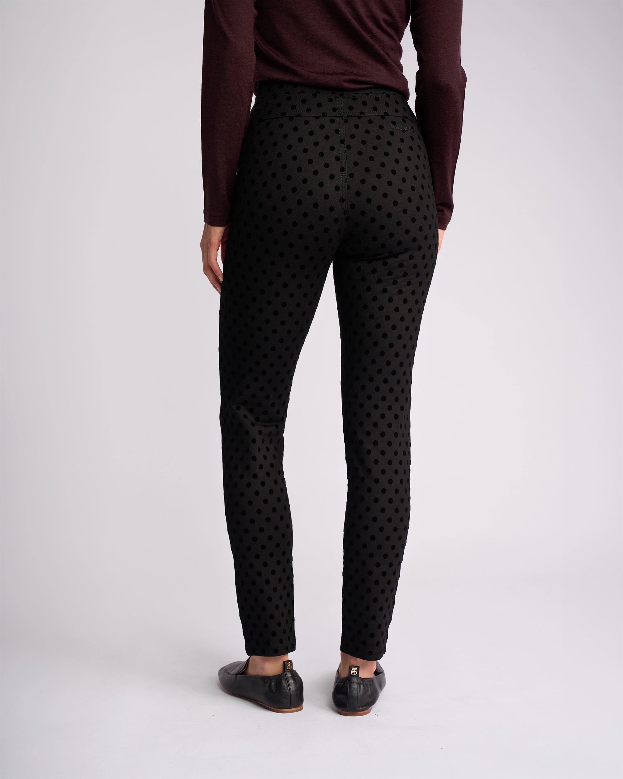 A person stands facing away, wearing the Vassalli View Pant - Black, paired with black shoes and a long-sleeve burgundy top against a plain light background.