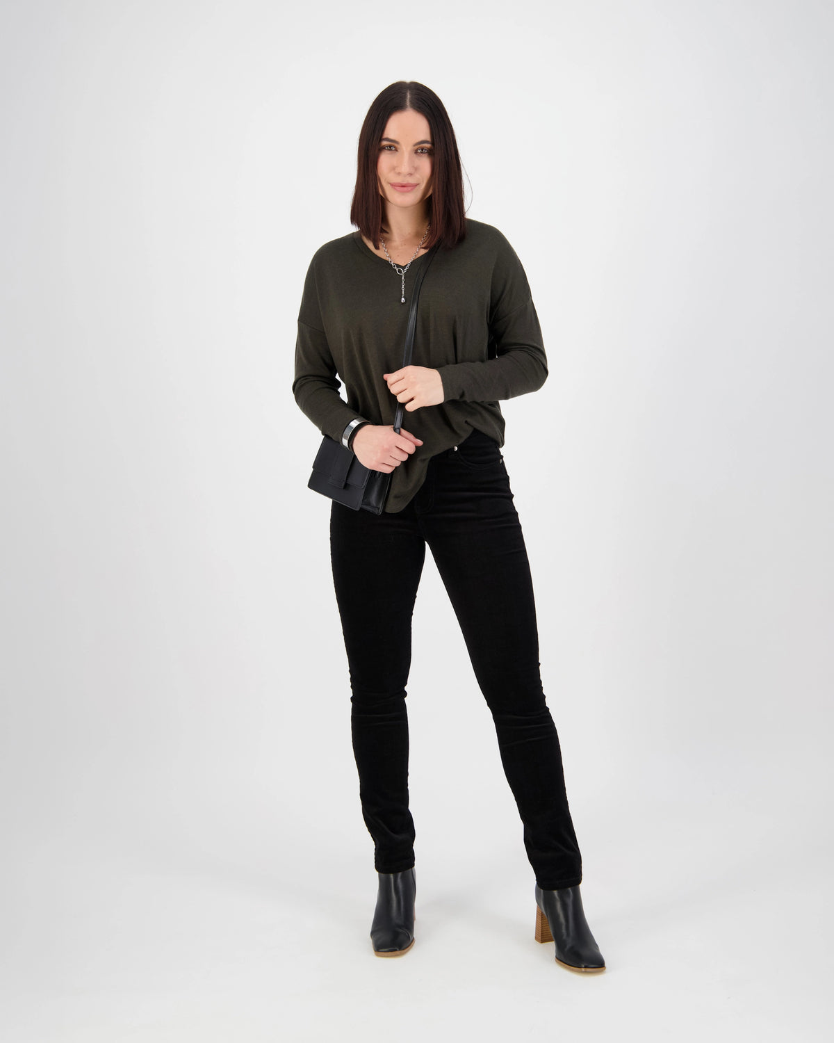 A woman with straight brown hair wears a dark green zippered top, Vassalli Park Cord Pant in black, black heeled boots, and holds a small black purse against a plain white background.