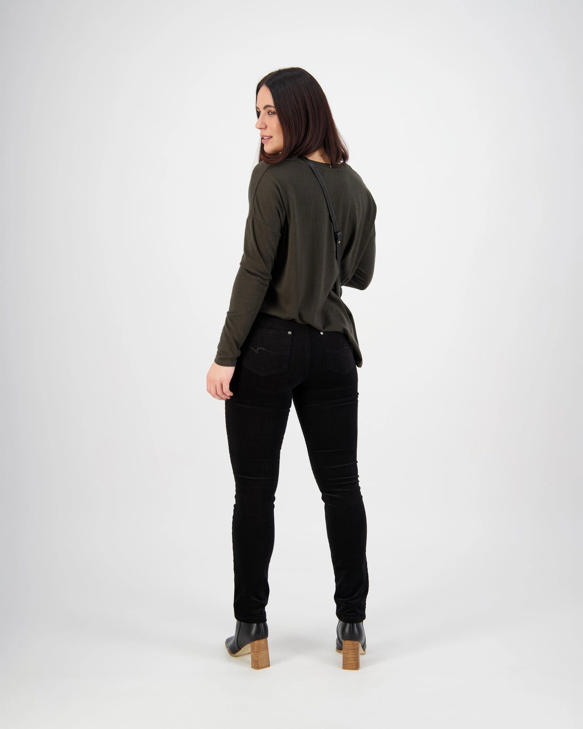 A woman with long dark hair models the Vassalli Park Cord Pant in Black, paired with a dark green top and black boots, standing against a plain white background and looking back over her shoulder.