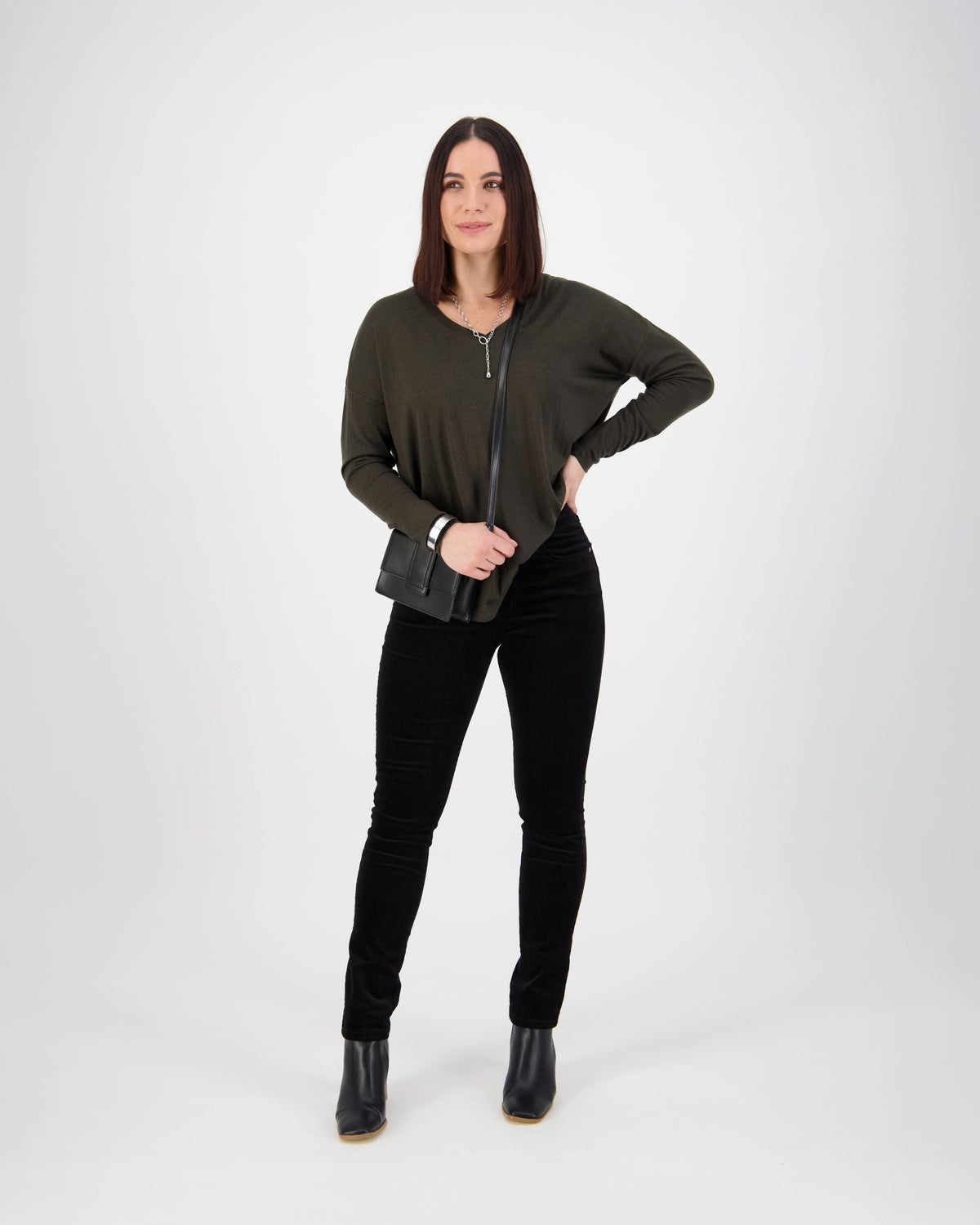 A woman with straight, shoulder-length brown hair stands against a white background, smiling slightly with one hand on her hip. She wears the Vassalli Park Cord Pant in black, paired with a dark green top, black boots, and a small black crossbody bag.