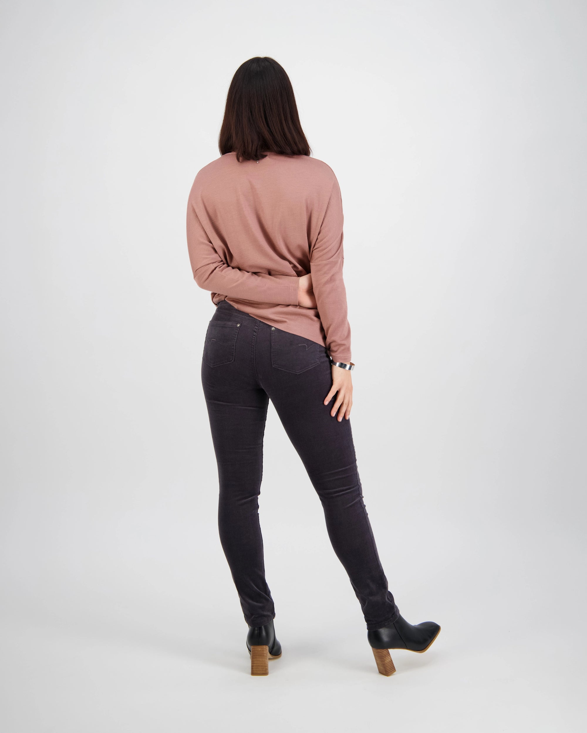 A person with shoulder-length dark hair, in a pink long-sleeve top and Vassalli Park Cord Pant - Charcoal, paired with black heeled boots, stands facing away against a plain white background.