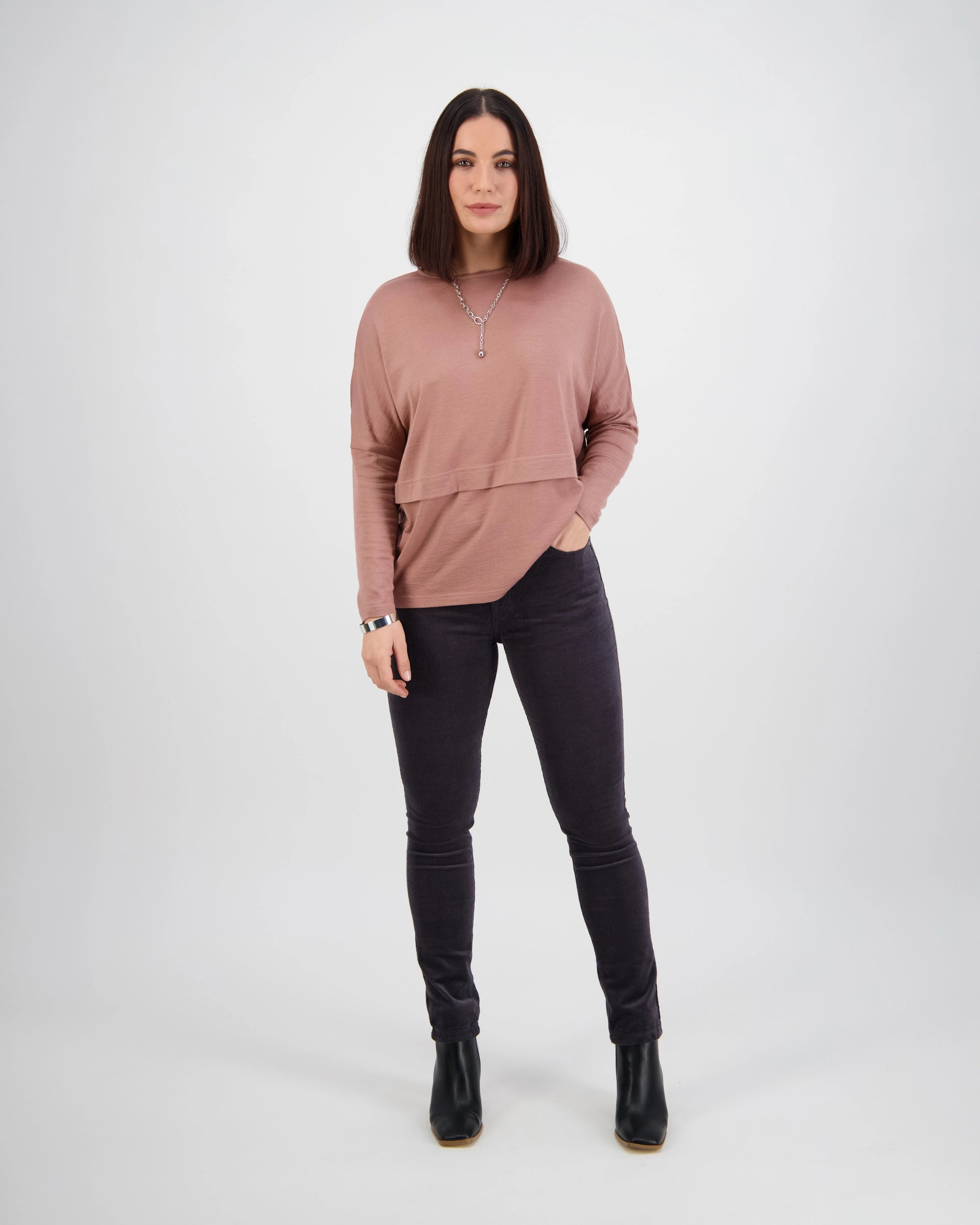 A woman stands against a plain white background, wearing Vassalli’s Park Cord Pant in Charcoal, a mauve long-sleeve top, black boots, and a necklace. Her hair is straight and parted in the middle, with one hand in her pocket.