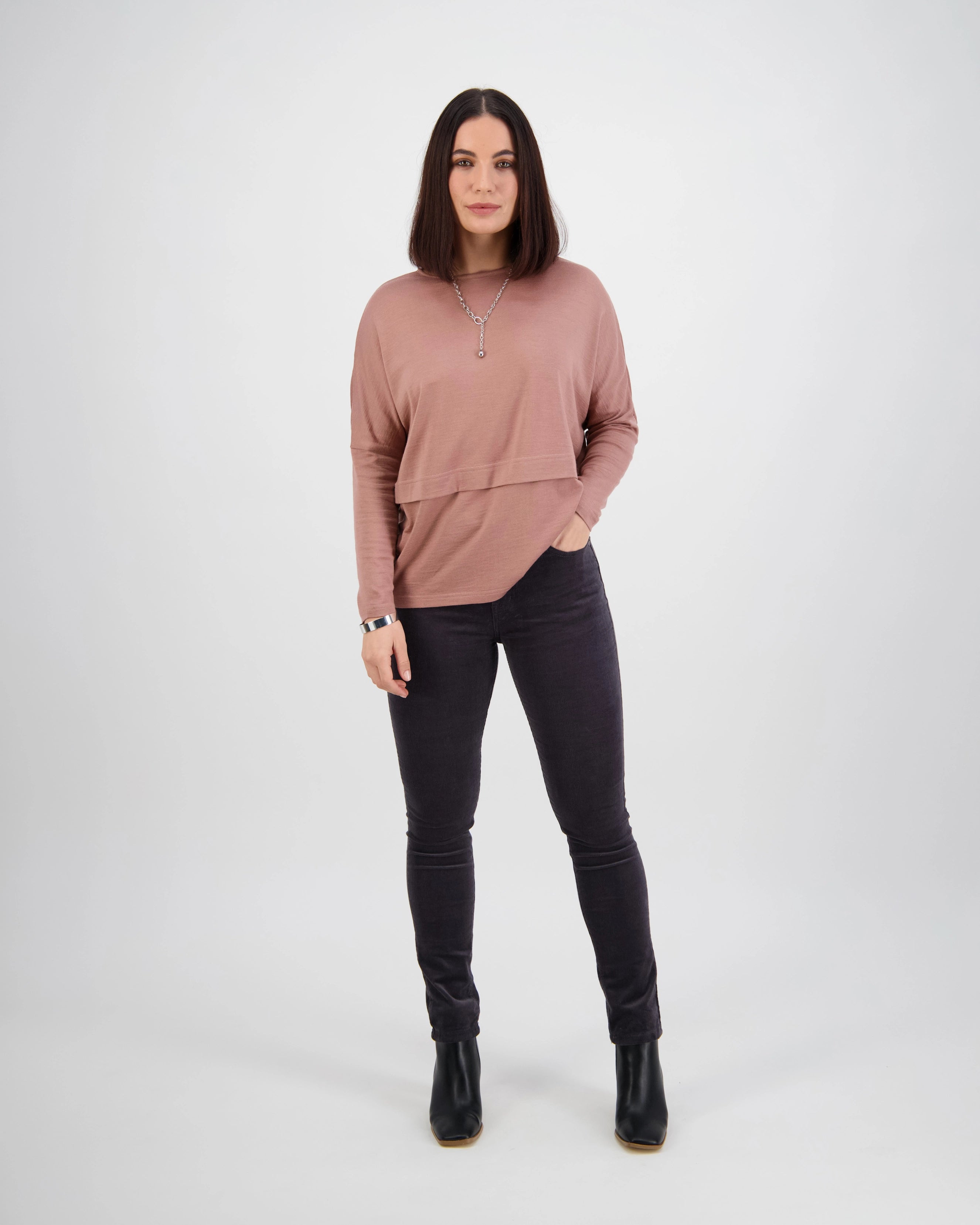 A woman stands against a plain white background, wearing Vassalli’s Park Cord Pant in Charcoal, a mauve long-sleeve top, black boots, and a necklace. Her hair is straight and parted in the middle, with one hand in her pocket.