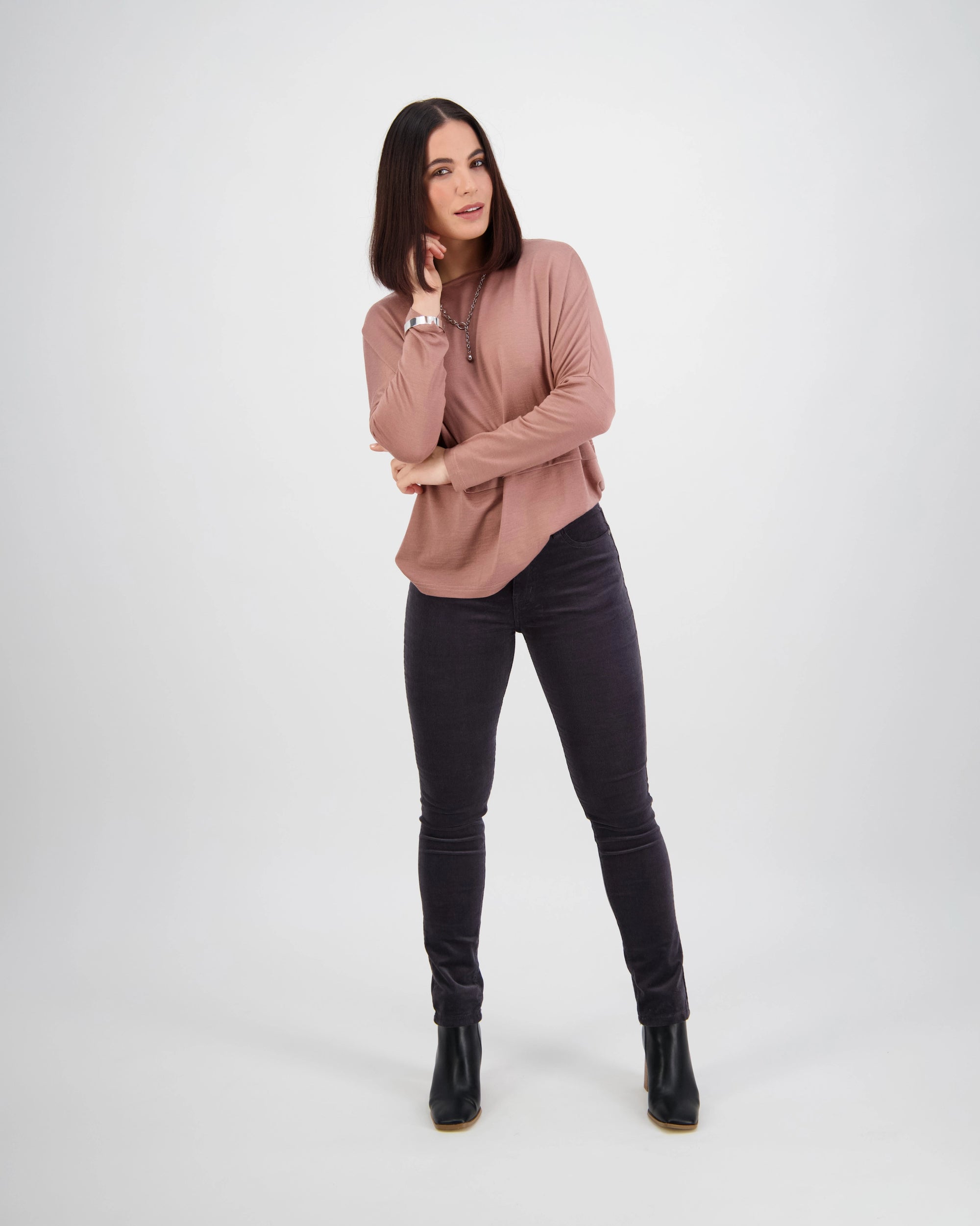 A woman with straight dark hair poses against a light background, wearing a mauve long-sleeve top, Vassalli Park Cord Pant in Charcoal, and black ankle boots, with one hand touching her face and the other at her side.