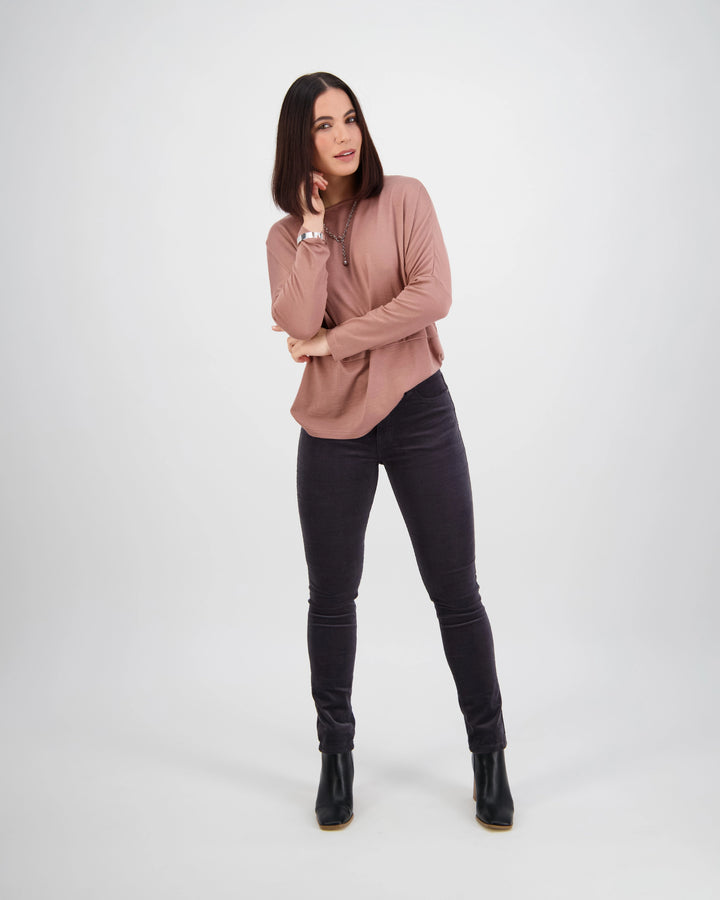 A woman with straight dark hair poses against a light background, wearing a mauve long-sleeve top, Vassalli Park Cord Pant in Charcoal, and black ankle boots, with one hand touching her face and the other at her side.