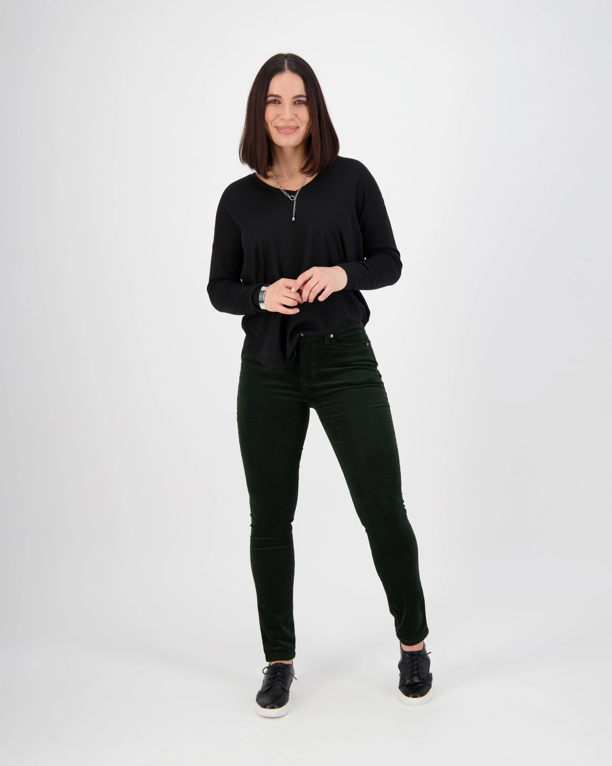 A woman with straight dark hair stands against a white background, smiling. She wears the Vassalli Park Cord Pant in Forest, a black long-sleeve top, and black shoes, with her hands clasped in front of her.