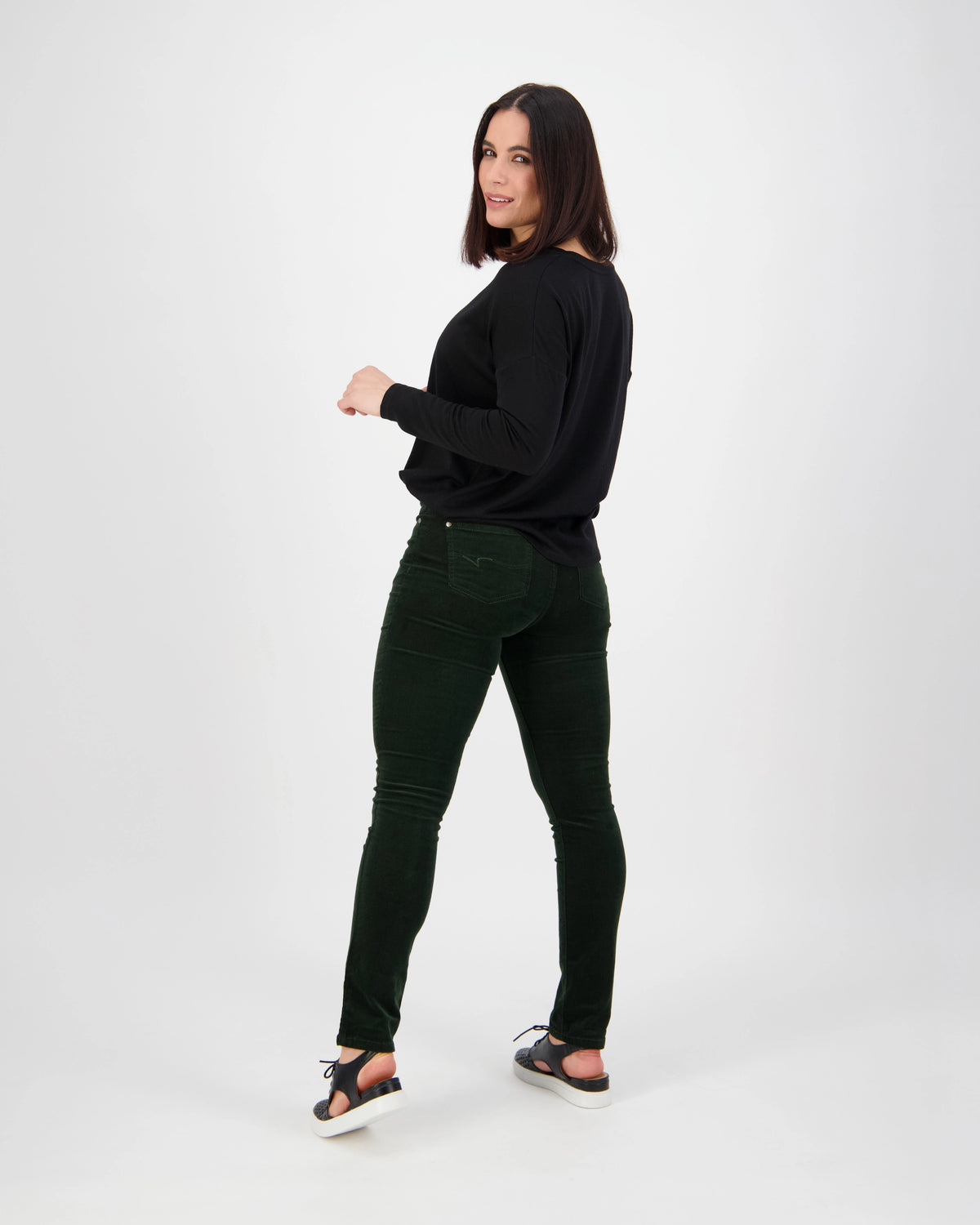 A woman with shoulder-length dark hair, wearing a black long-sleeve top, black sandals, and Vassalli’s Park Cord Pant in Forest, stands against a white background looking over her shoulder and smiling.