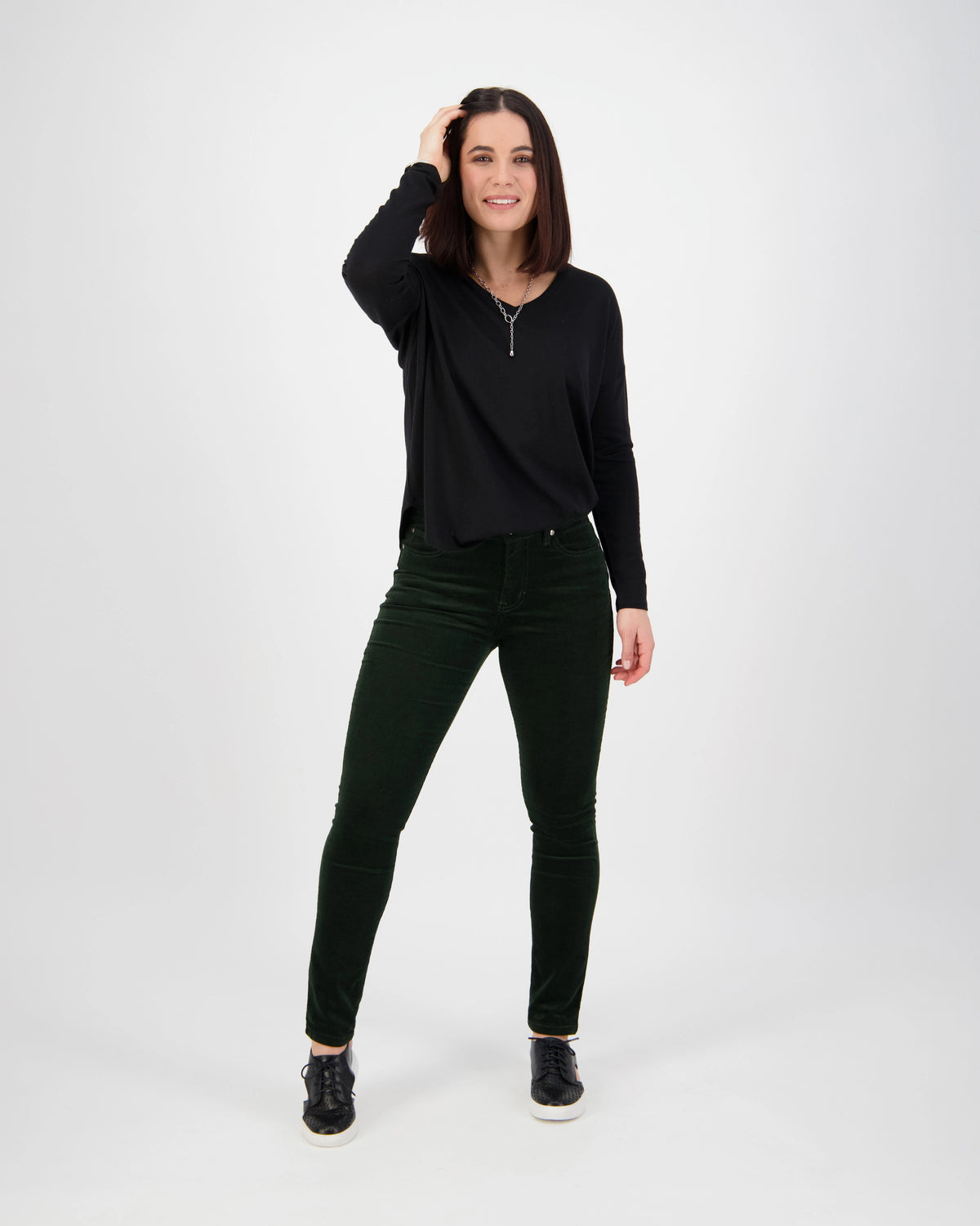 A woman with straight dark hair smiles in a relaxed pose against a white background, wearing the Vassalli Park Cord Pant in Forest, a black long-sleeve top, and black shoes, with one hand touching her hair.