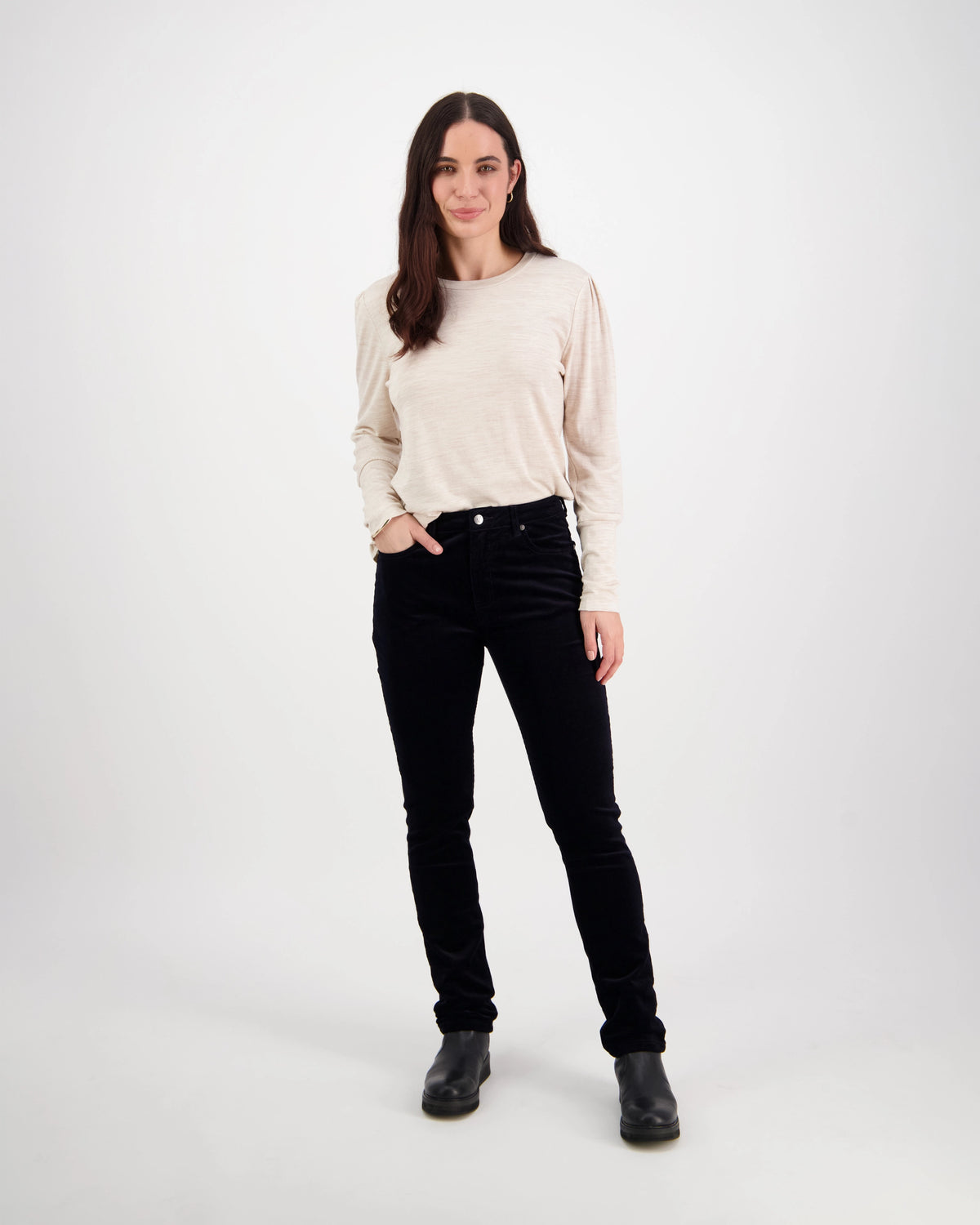 A woman with long brown hair stands against a white background, wearing a beige long-sleeve top and Vassalli Park Cord Pant in Midnight. She has one hand in her pocket and smiles gently at the camera.