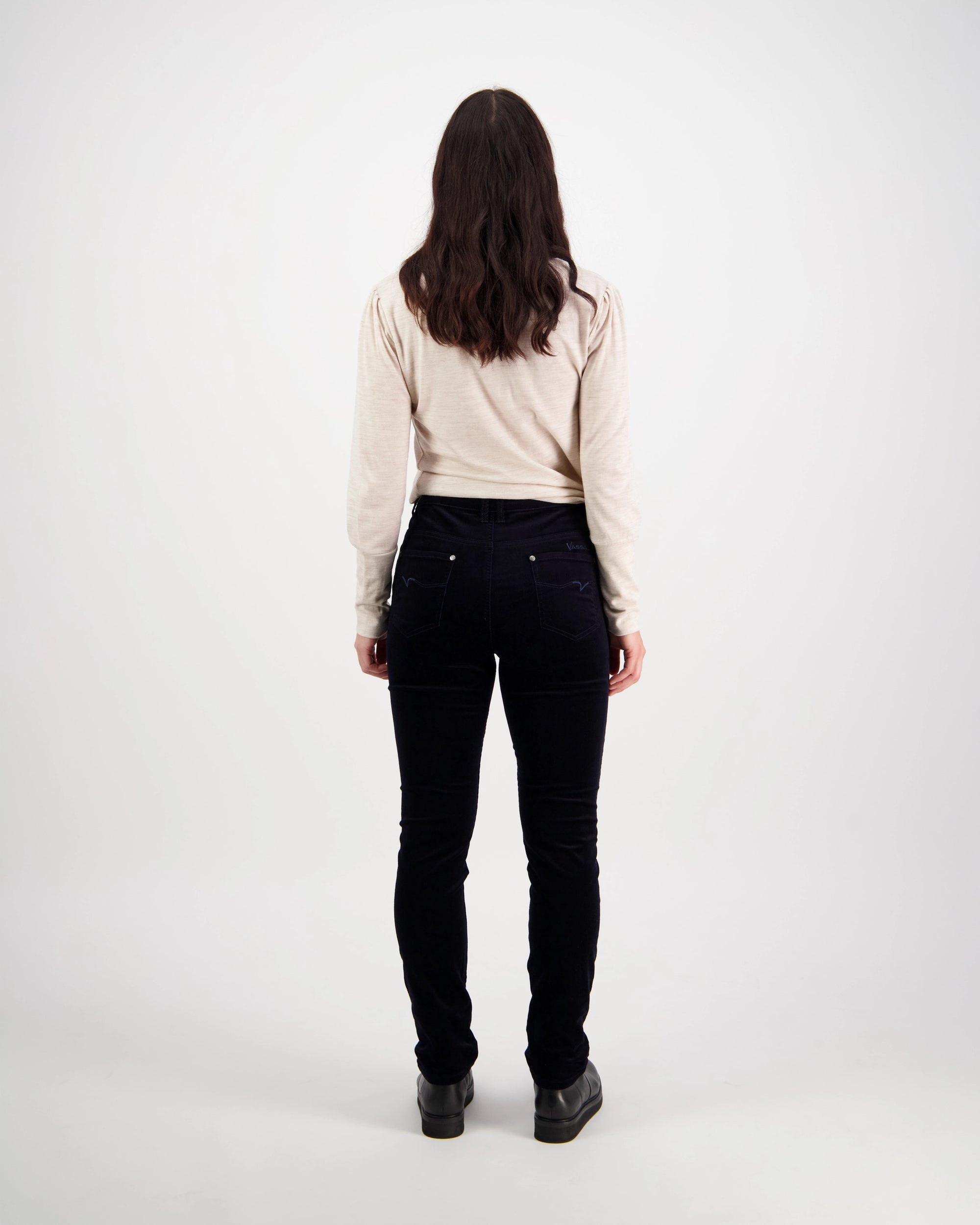 A person with long brown hair, wearing a beige long-sleeve top and Vassalli Park Cord Pant in Midnight, stands facing away from the camera against a plain white background.