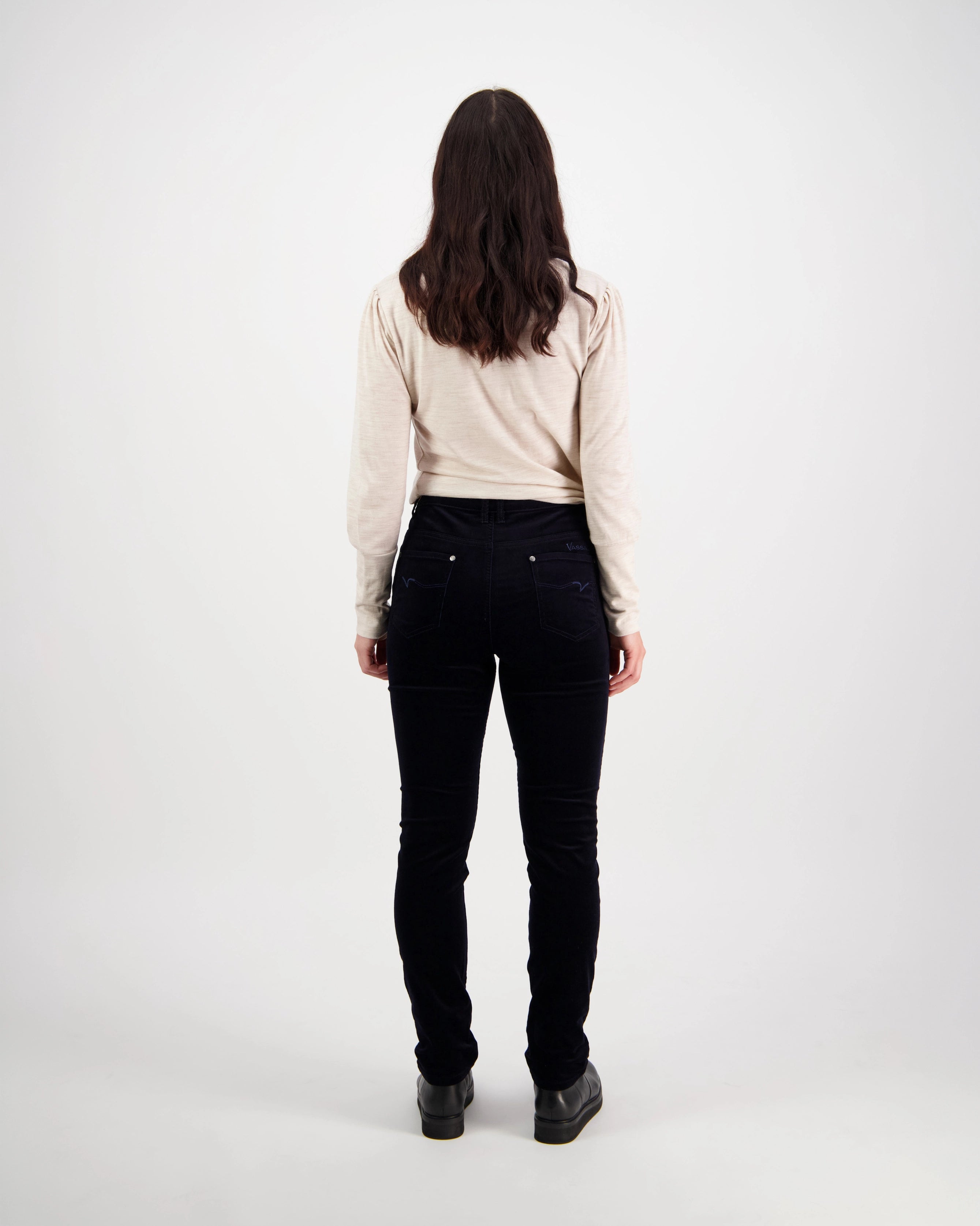 A person with long brown hair, wearing a beige long-sleeve top and Vassalli Park Cord Pant in Midnight, stands facing away from the camera against a plain white background.