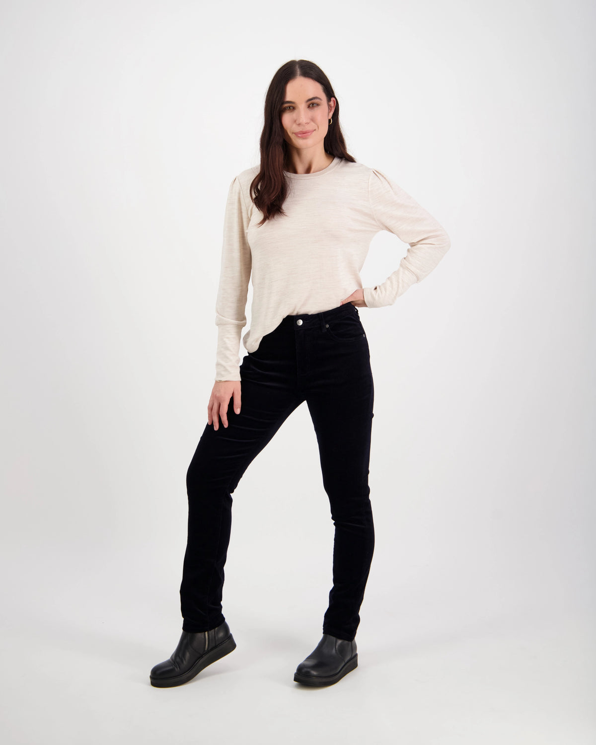 A woman with long dark hair stands confidently against a white background, wearing a cream long-sleeve top, black boots, and the Vassalli Park Cord Pant in Midnight, one hand in her pocket and a slight smile.