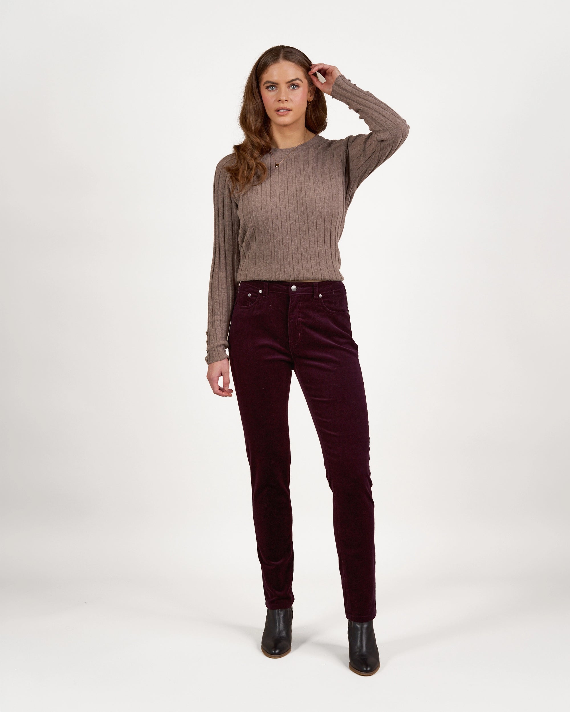 A woman faces forward against a white background, wearing a taupe ribbed sweater, Vassalli Park Cord Pant in Mulberry, and black ankle boots. One arm is bent near her temple, and she has long wavy brown hair.