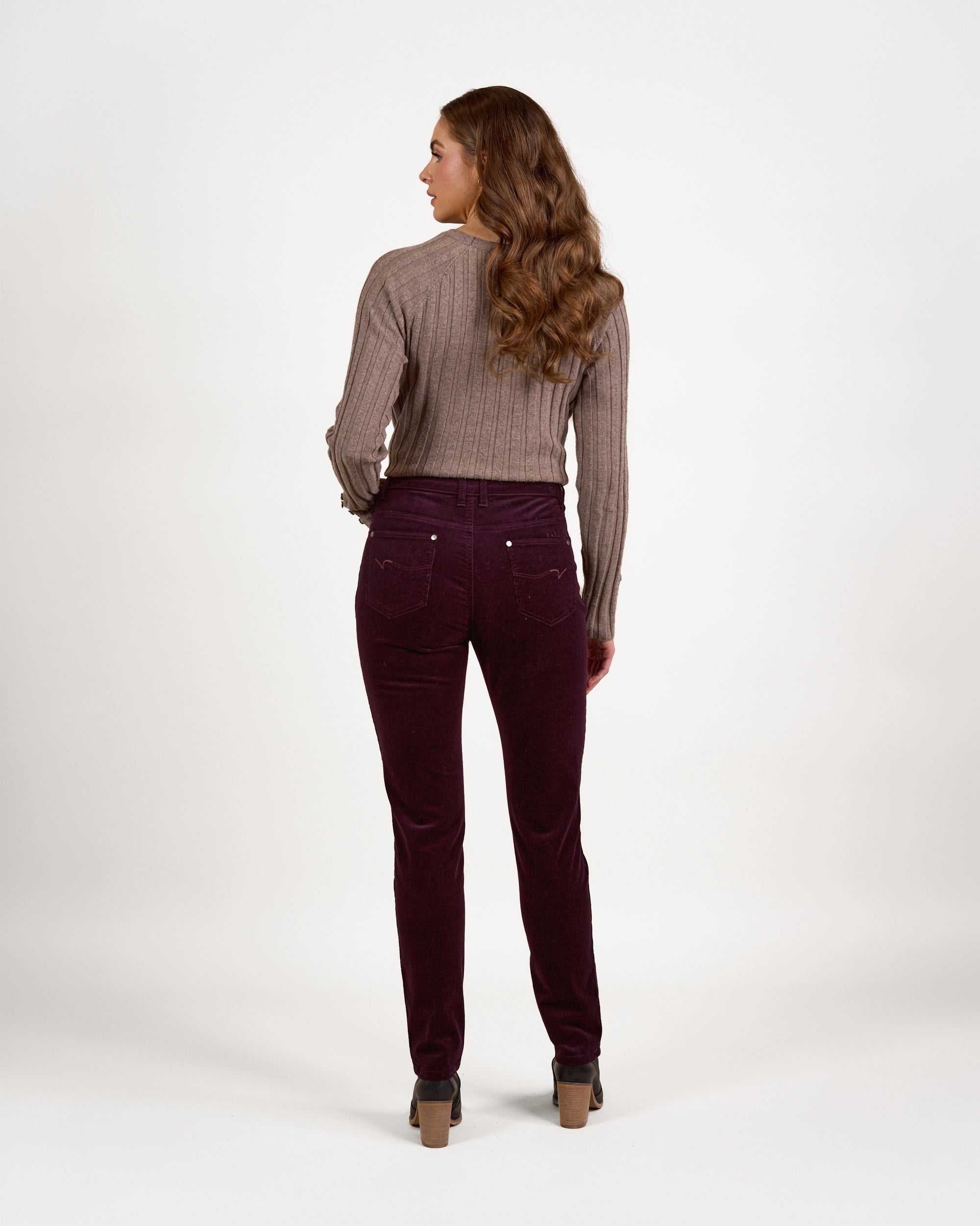 A woman with long wavy hair faces away, wearing a brown ribbed sweater, Vassalli’s Park Cord Pant in Mulberry, and beige heeled shoes, set against a plain white background.