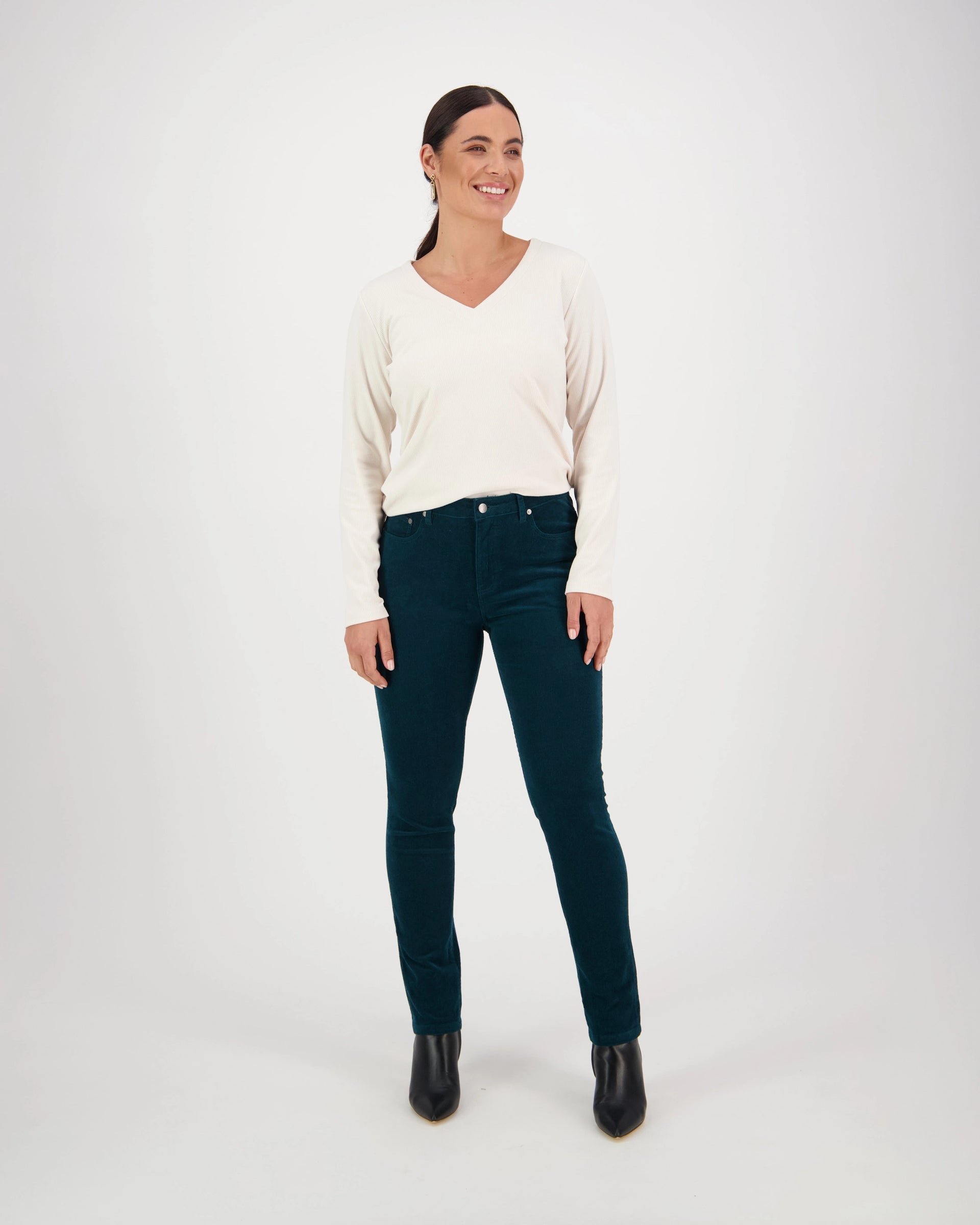 A woman smiles against a white background, wearing a cream long-sleeve V-neck sweater, black ankle boots, and Vassalli Park Cord Pant in Peacock.
